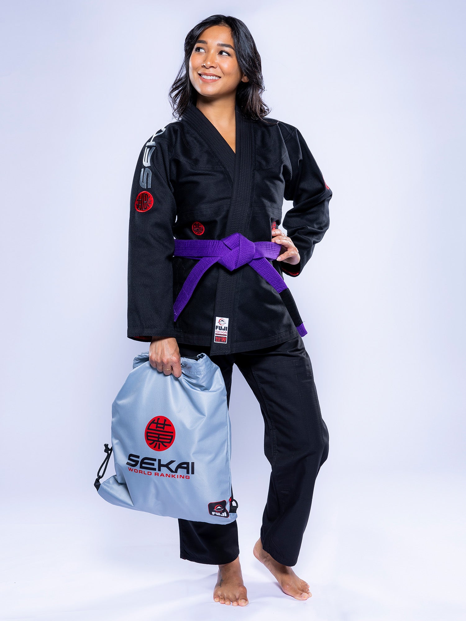 A woman wearing the Womens SEKAI 3.0 BJJ Gi Black Grey and a purple belt stands barefoot, smiling and holding a gray gym bag with SEKAI World Hanegin branding.