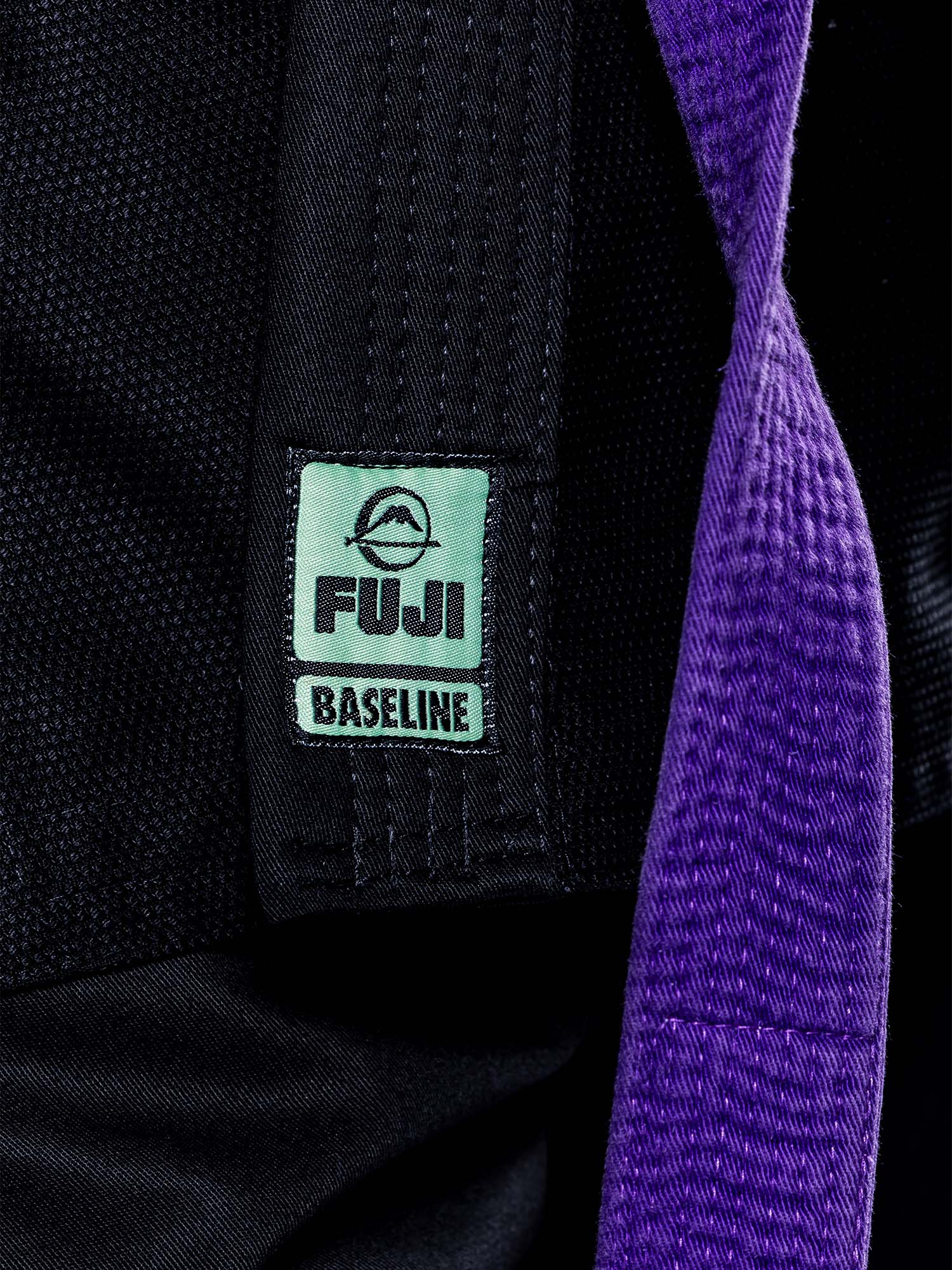 Close-up of the Womens Baseline 2.0 Slim Fit BJJ Gi in Black Mint, featuring a green FUJI Baseline label on black fabric with a textured purple strap draped across it.