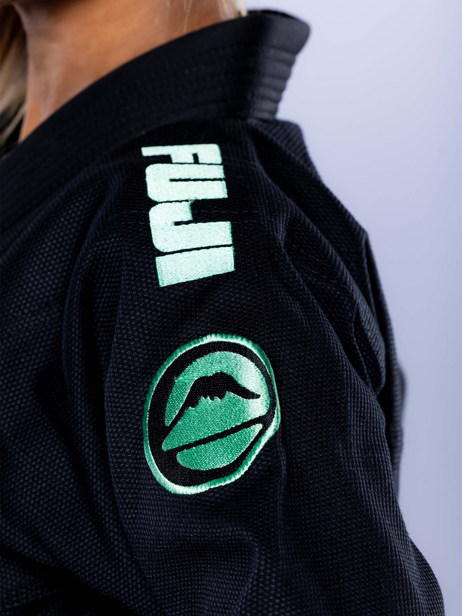 Close-up of a person wearing the Womens Baseline 2.0 Slim Fit BJJ Gi in black mint with FUJI text and a green mountain logo embroidered on the shoulder.