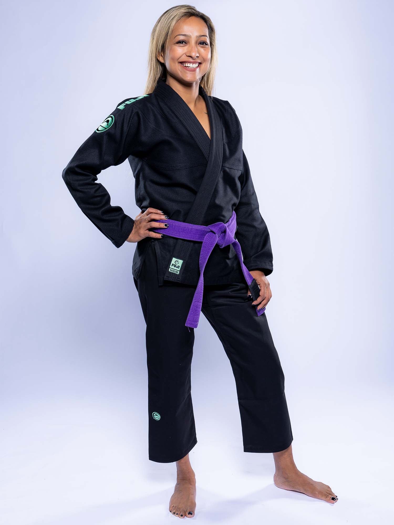 A blonde woman stands barefoot on a white background, smiling confidently in the Womens Baseline 2.0 Slim Fit BJJ Gi Black Mint with a purple belt. Her left hand rests on her hip as she poses relaxed and proud.