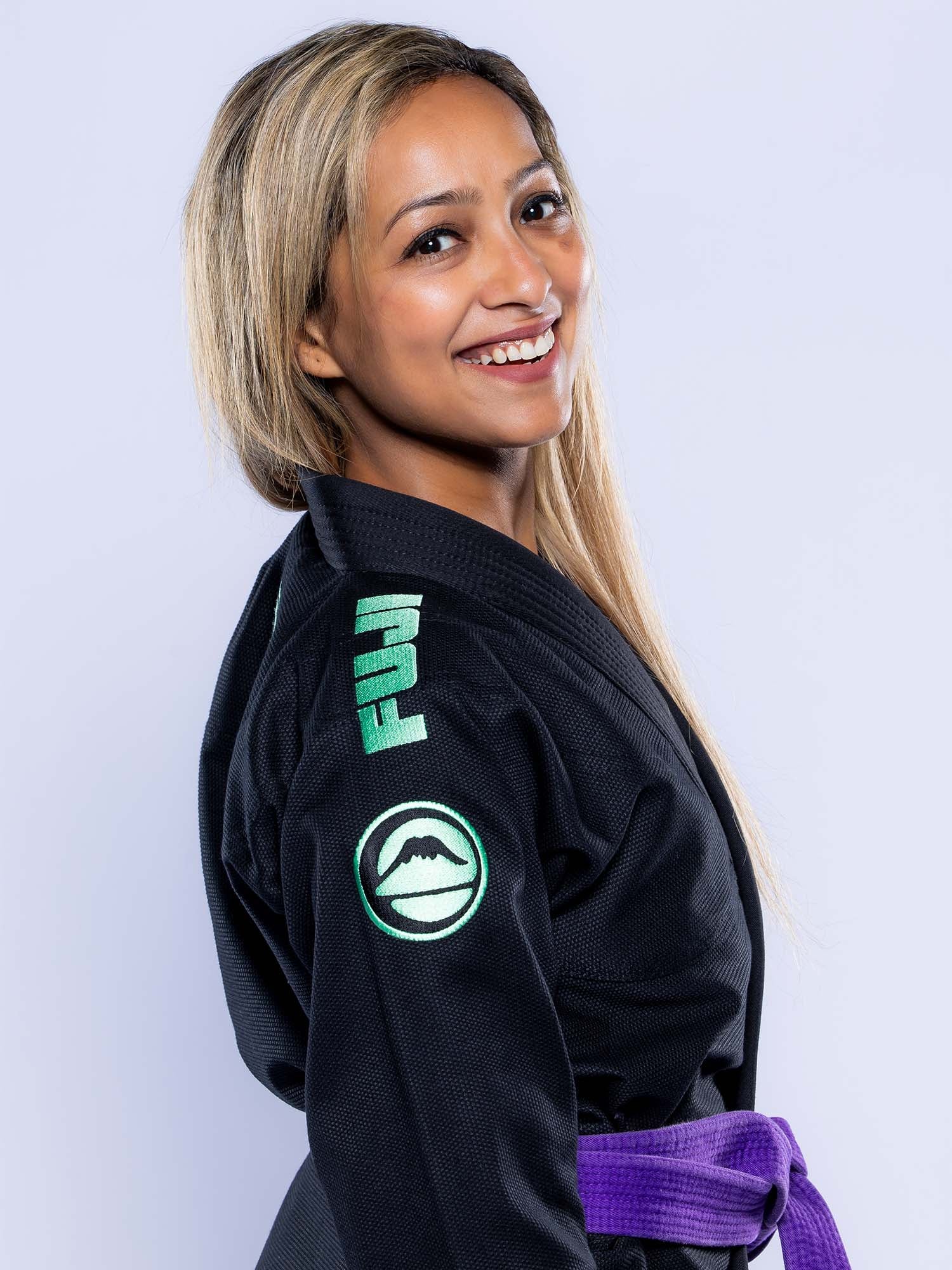 A woman with long, light brown hair smiles while wearing the Womens Baseline 2.0 Slim Fit BJJ Gi Black Mint, featuring mint green patches and a purple belt, standing against a light background.