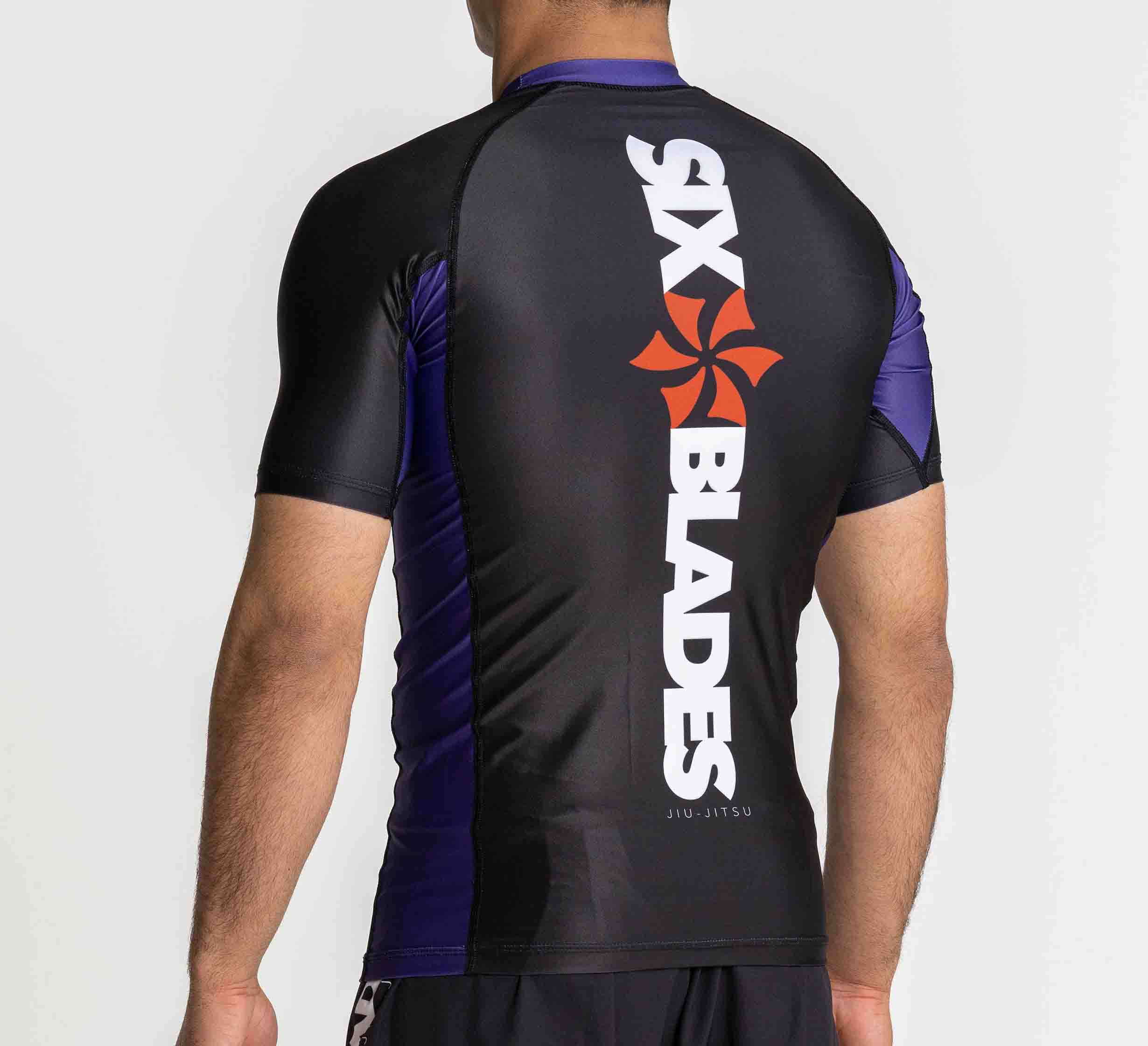 A person wearing the Six Blades Short Sleeve Rashguard Purple, featuring black and purple panels and an orange pinwheel logo with "SIX BLADES" printed vertically on the back, stands facing away from the camera.