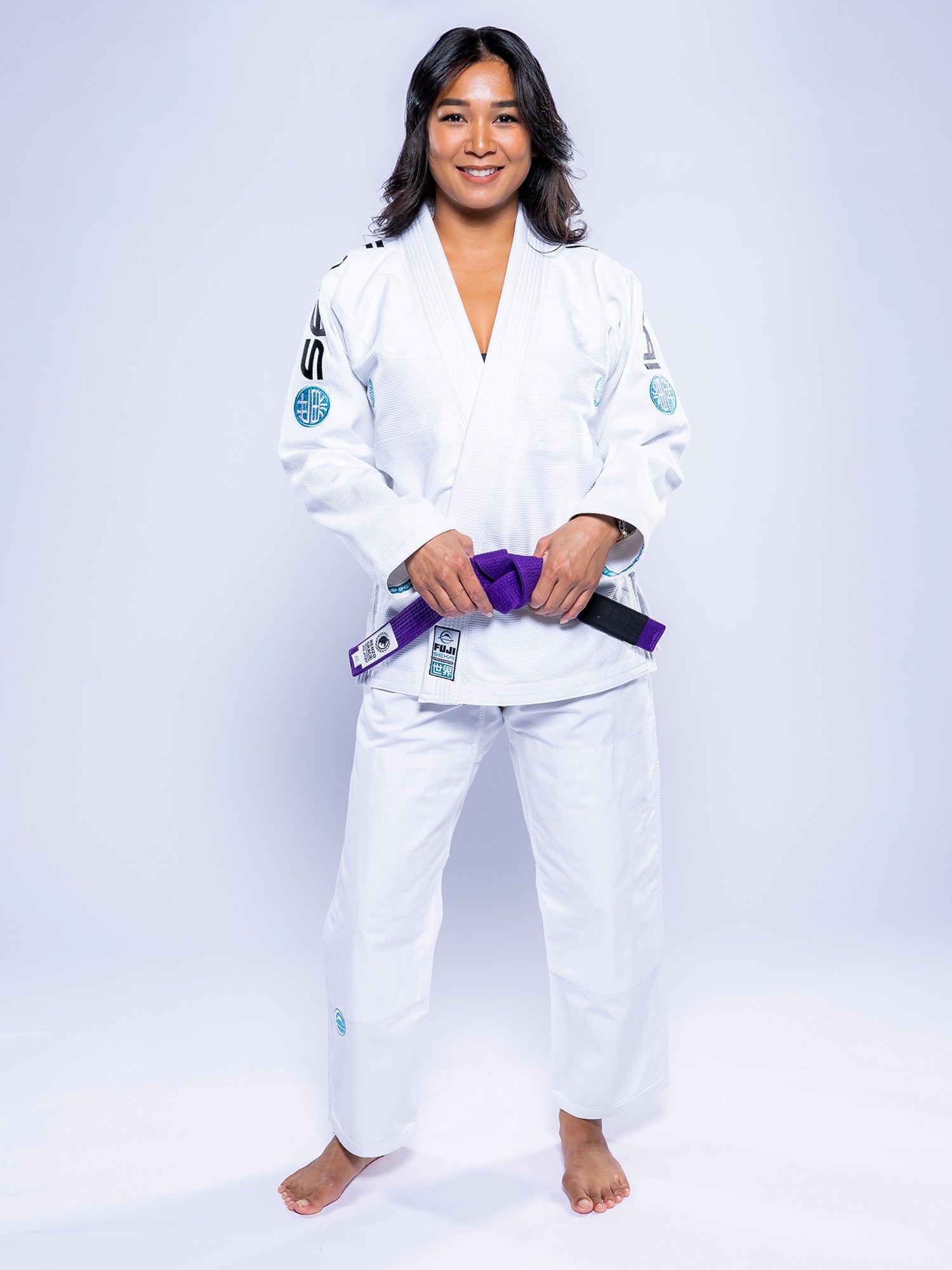 A woman stands barefoot on a white background, smiling and wearing the Womens SEKAI 3.0 BJJ Gi White Teal with a purple belt, which she holds in front of her.
