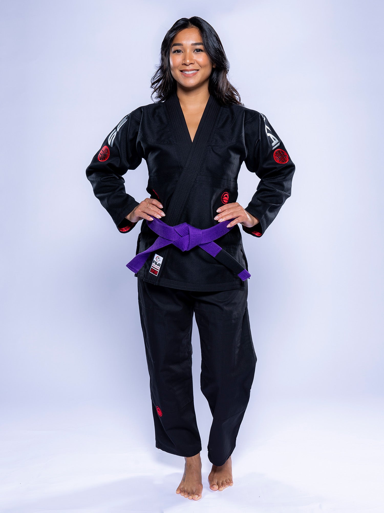 A woman with long dark hair stands barefoot, smiling with hands on hips, wearing the Womens SEKAI 3.0 BJJ Gi in black grey with a purple belt, against a light background.