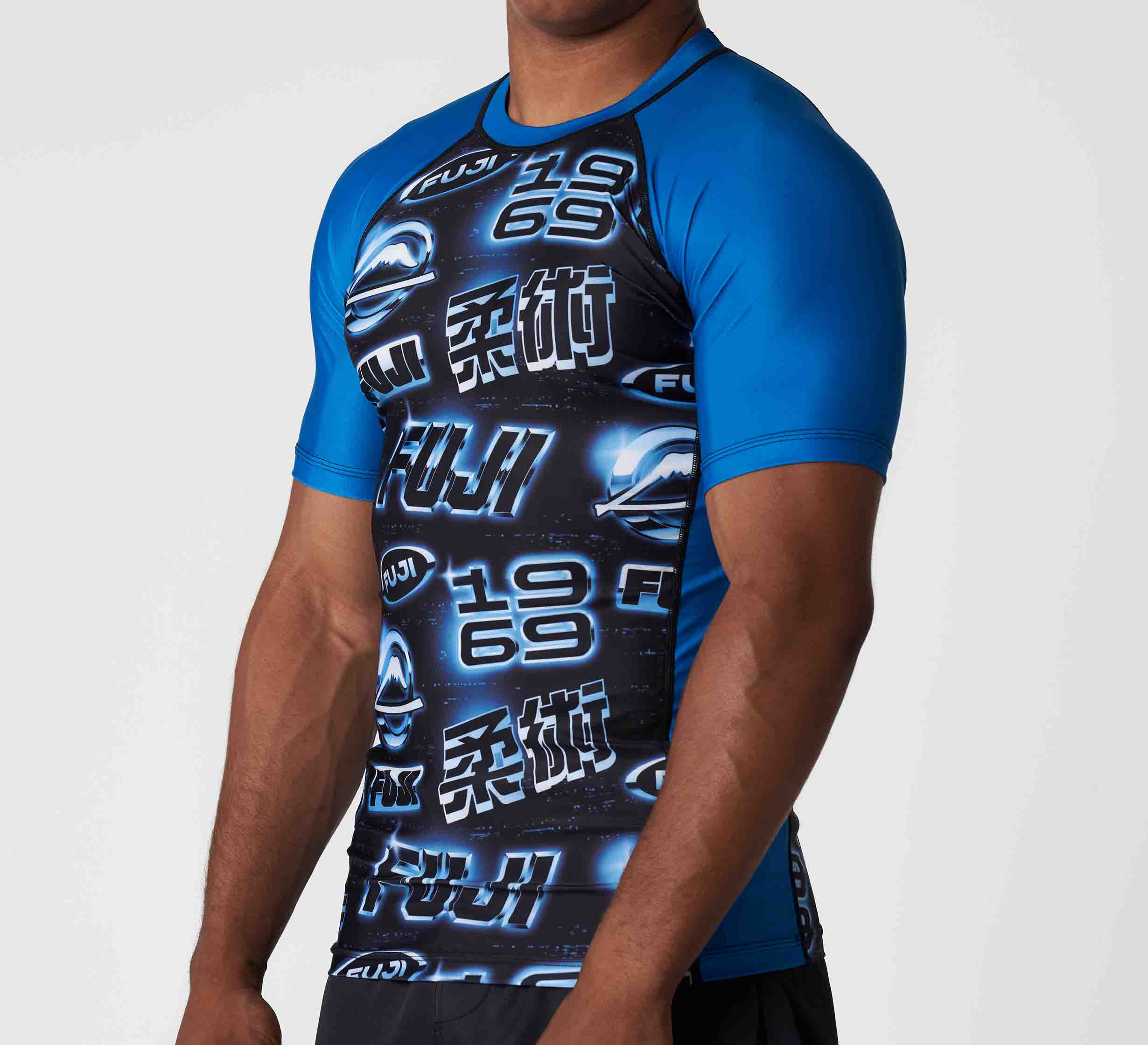 Someone is wearing the Synth Flex Lite Rashguard Blue, a blue and black short-sleeve athletic shirt with logos, numbers, and Japanese characters on the front, standing against a plain white background.