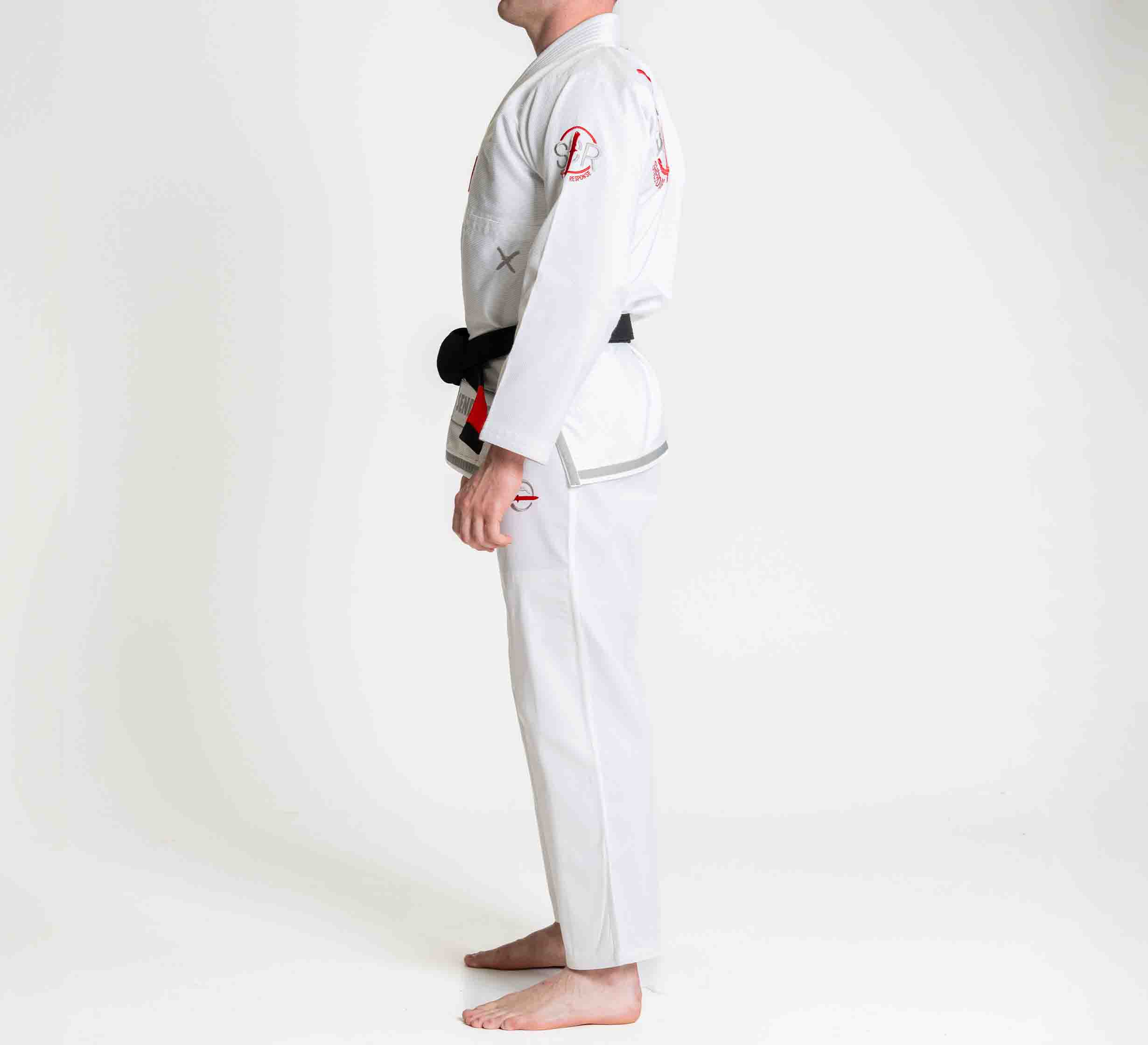 A person stands barefoot in profile, wearing the Suparaito BJJ Gi Sheepdog Response Edition White with patches and a black belt, against a plain white background.