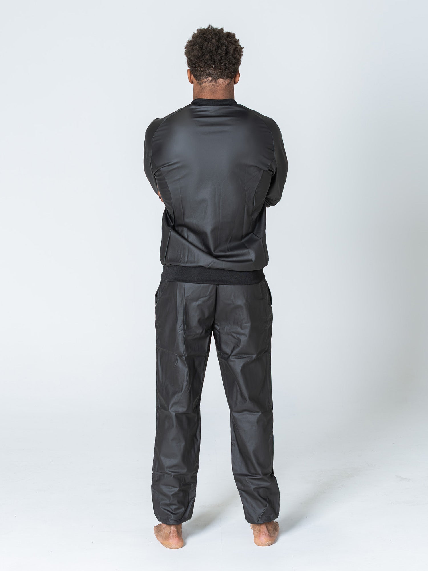 A man stands barefoot on a white background, facing away from the camera with arms crossed, wearing the Grappling Sauna Suit Black.