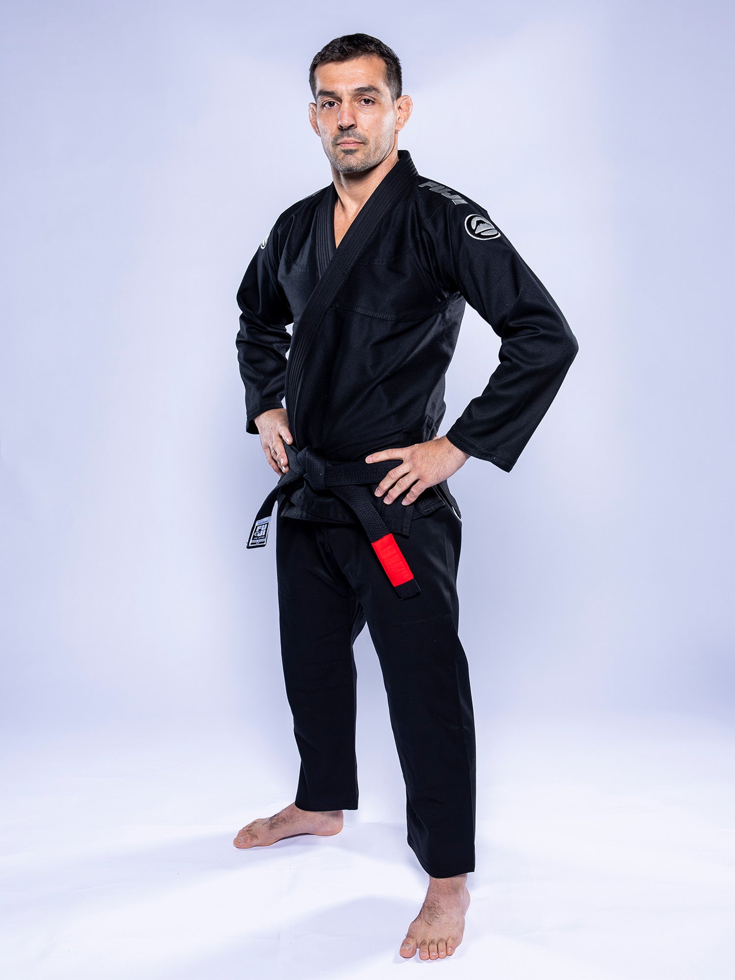 A man stands confidently with hands on hips, barefoot on a white background, wearing the Baseline 2.0 BJJ Gi Black Grey and a black belt with a red bar.