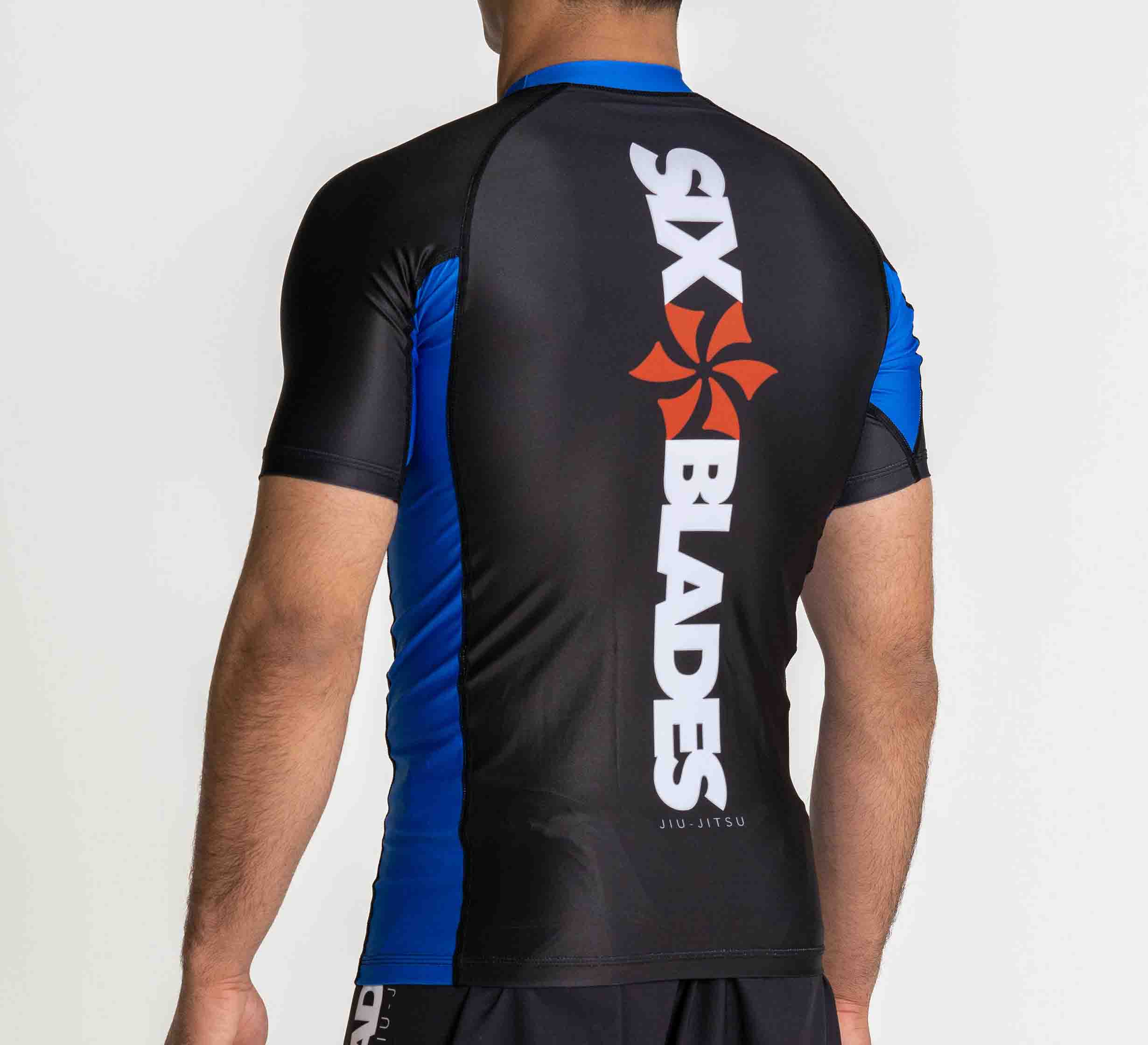 A man is seen from behind wearing the Six Blades Short Sleeve Rashguard Blue, featuring black and blue colors with "SIX BLADES" and an orange logo printed vertically on the back.