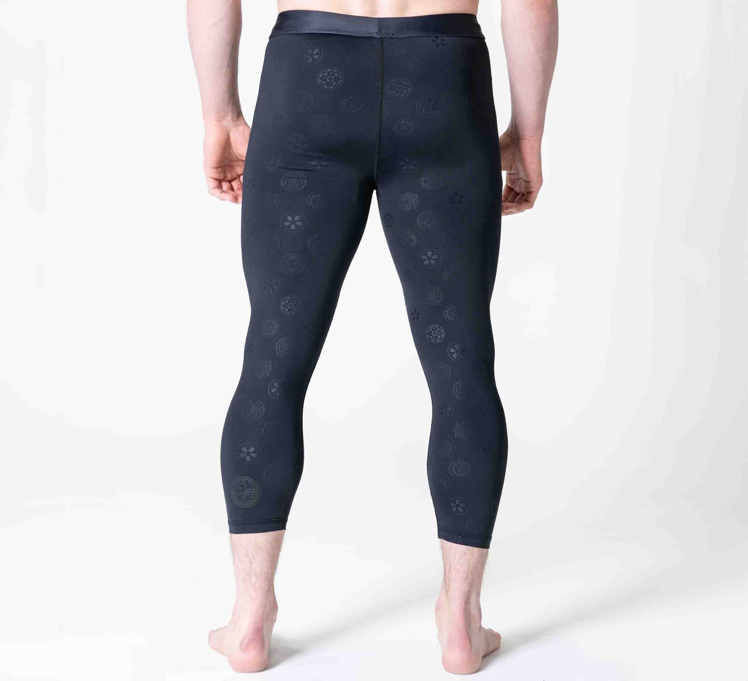 A person stands barefoot on a white surface, facing away from the camera, wearing Shogun Heat Gear Spats Black—fitted athletic leggings with a subtle circular pattern.