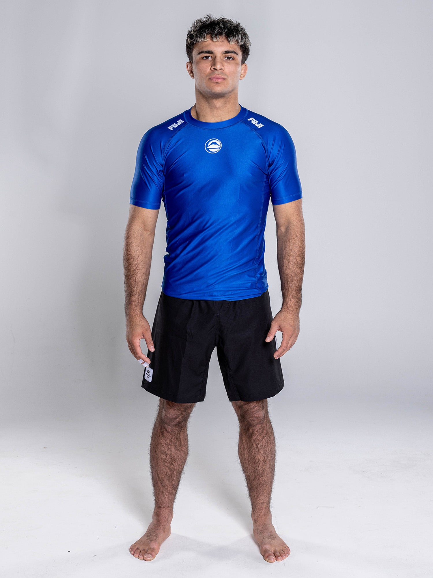 A person with short curly hair stands barefoot on a white background, facing forward with arms relaxed, wearing the Baseline 2.0 Short Sleeve Rashguard Navy and black shorts.