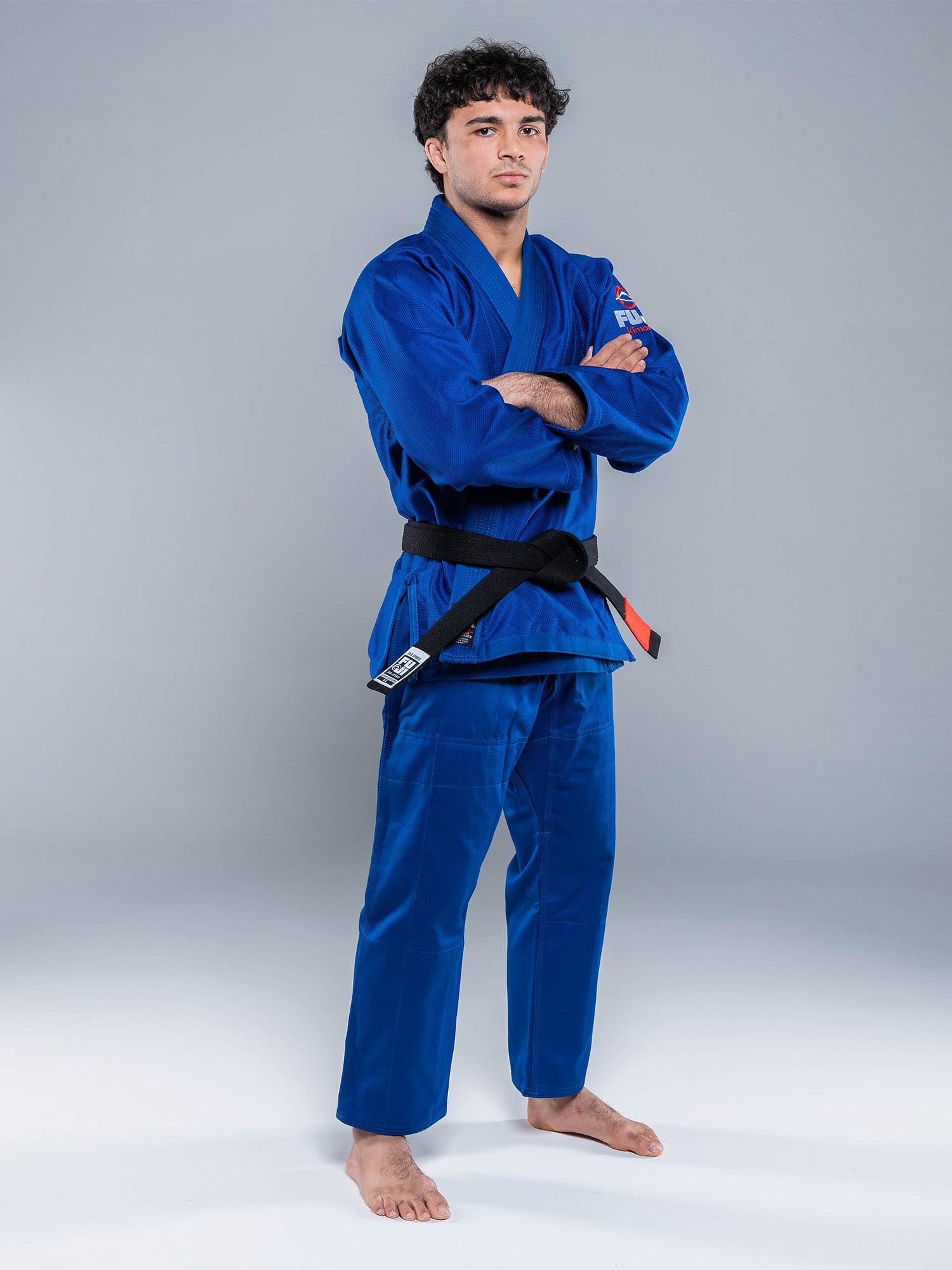 A man confidently poses barefoot on a plain background, arms crossed, wearing the Lightweight BJJ Gi Blue and a black belt.