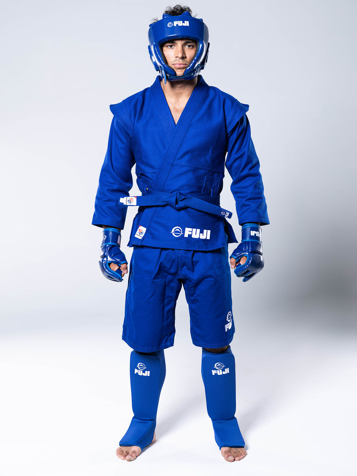 A person wearing a blue FIAS Sambo Kurtka Jacket Blue With Blue Belt, shorts, protective headgear, gloves, and shin guards.