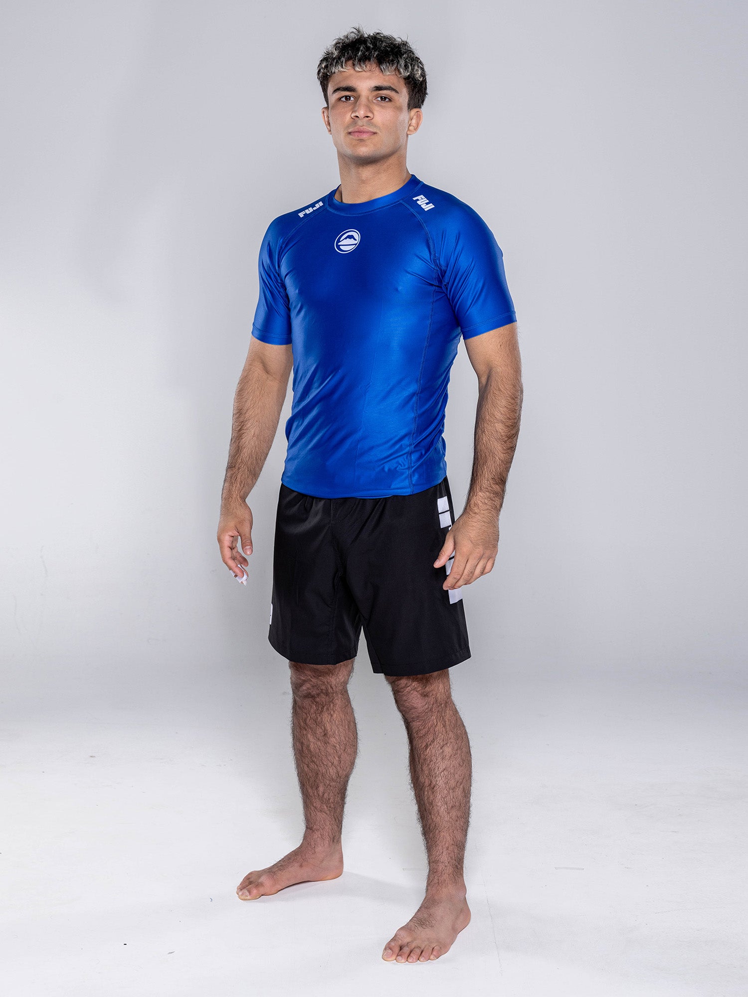 A young man stands barefoot on a white background, wearing the Baseline 2.0 Short Sleeve Rashguard Navy and black shorts, arms relaxed at his sides, looking straight ahead.