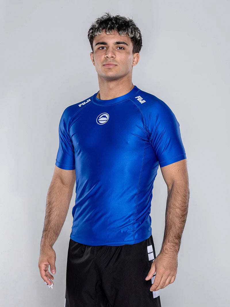 A young man with short, dark hair and subtle highlights wears the Baseline 2.0 Short Sleeve Rashguard Blue, paired with black shorts, standing against a plain light background and facing the camera with a neutral expression.