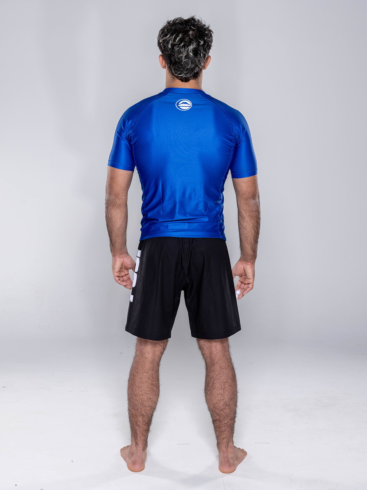A man stands barefoot facing away from the camera, wearing the Baseline 2.0 Short Sleeve Rashguard Navy and black shorts against a plain light gray background.