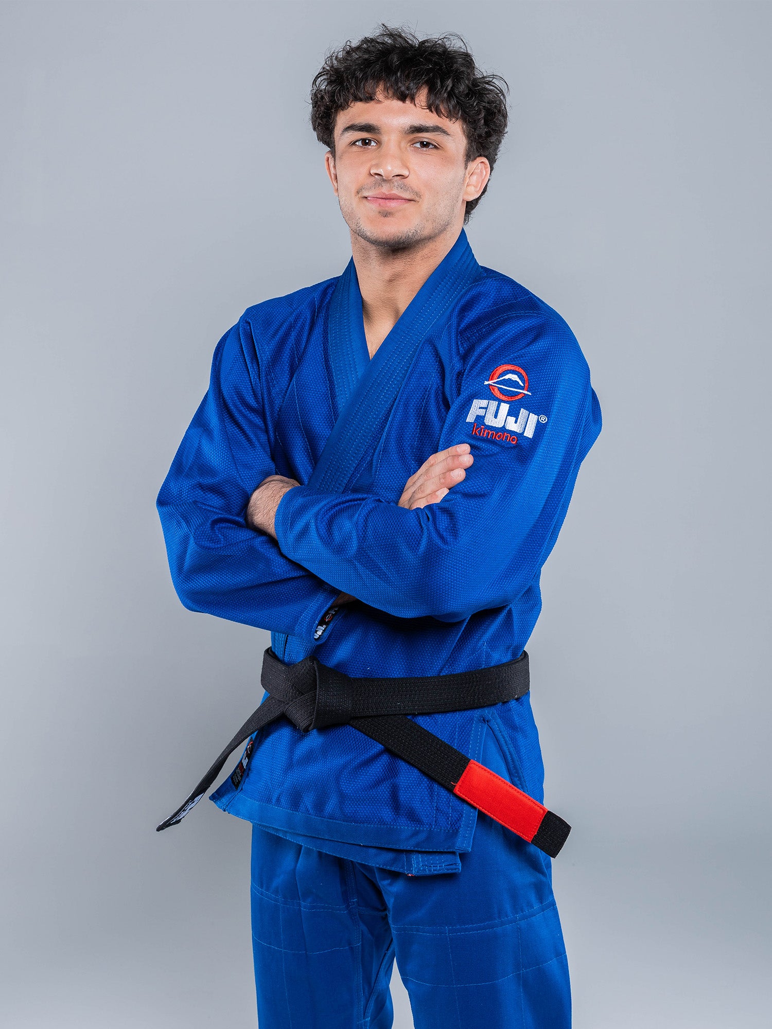 A young man wearing the Lightweight BJJ Gi Blue with a black belt stands, arms crossed, smiling slightly at the camera. The gi features a Fuji logo on the sleeve and is set against a plain light gray background.
