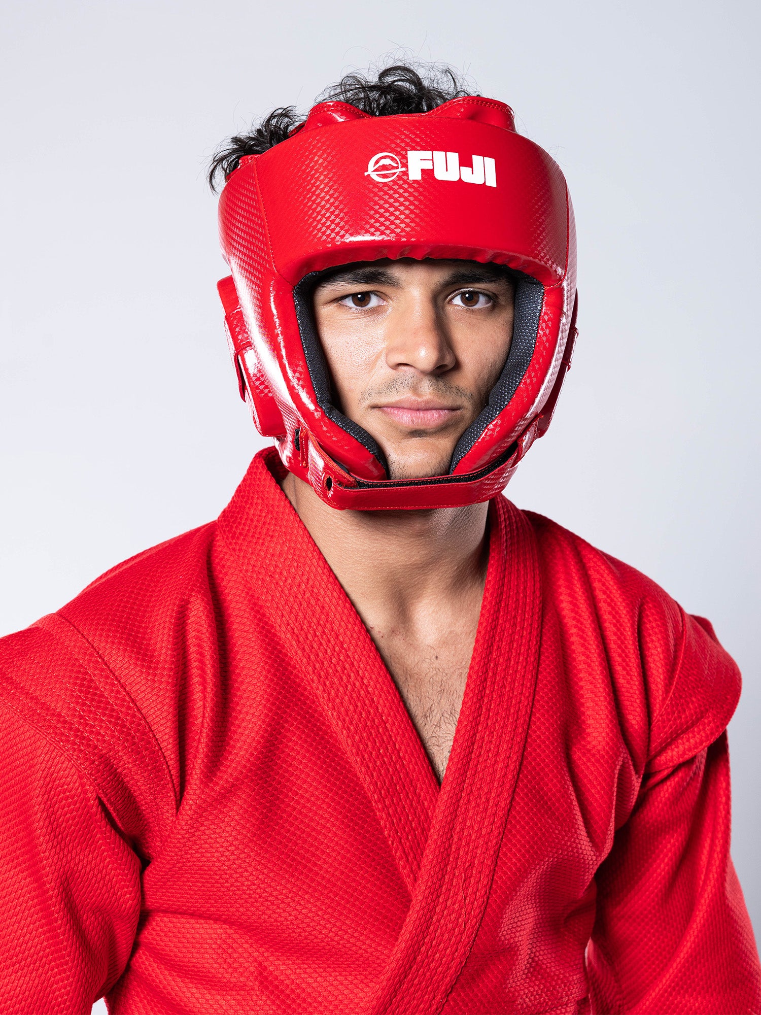 A young man with dark hair wears a bright red FIAS Sambo Combat Headgear and a matching red sambo uniform, looking directly at the camera against a plain, light grey background.