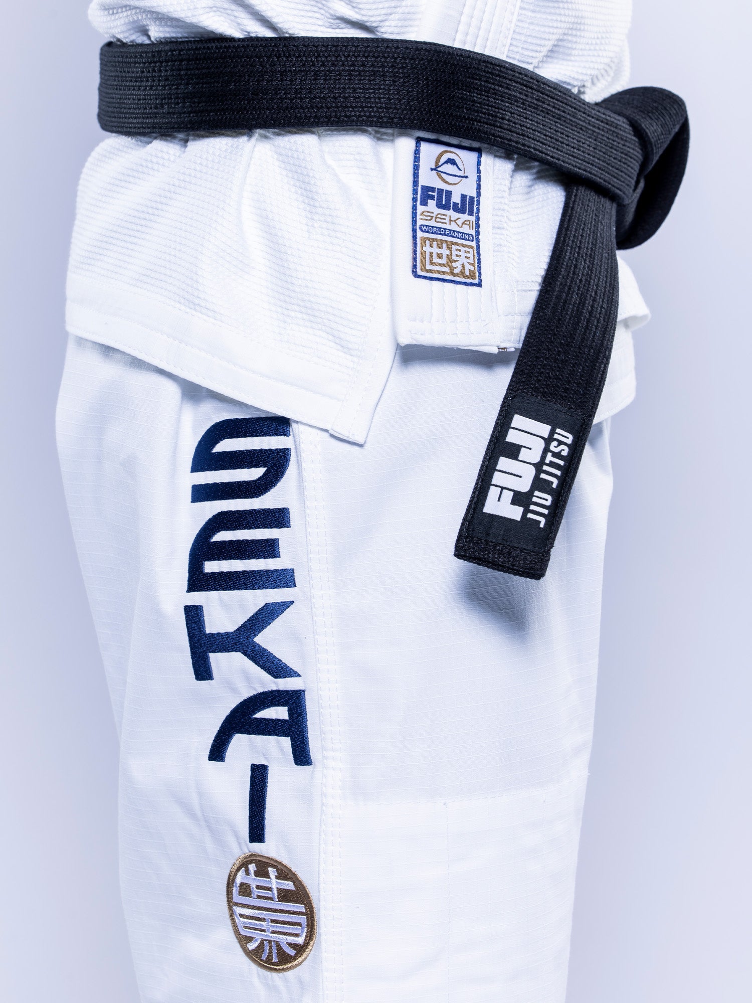 Close-up of a person wearing the SEKAI 3.0 BJJ Gi White Navy with blue Sekai lettering, a black belt, and Fuji brand patches on both the gi and the belt.