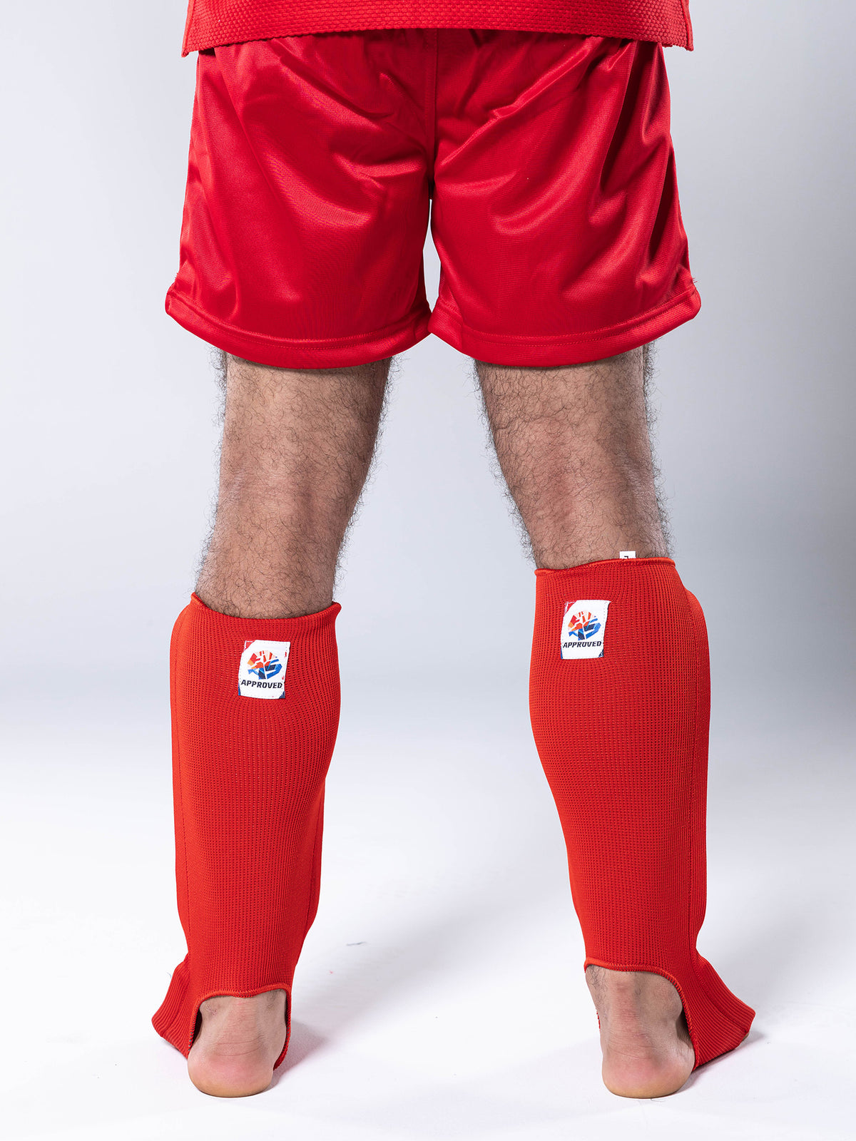 Person wearing FIAS Sambo Combat Shin Guards Red and red shorts, standing barefoot with their back facing the camera against a plain light background.