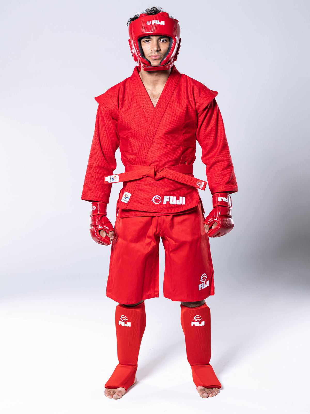 A person wearing a complete red FIAS Sambo Kurtka Jacket Red With Red Belt, including shoulder epaulets, shorts, headgear, fingerless gloves, and shin guards.