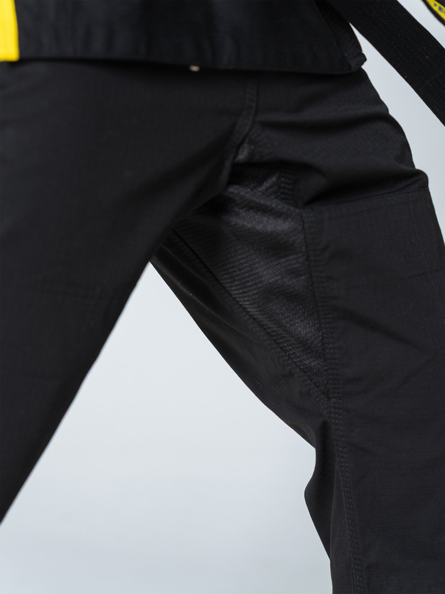 Close-up of a person wearing the Special Edition Holiday Flow Tech Jiu Jitsu Gi Black, highlighting the textured fabric and detailed stitching. The gi features black pants and a black belt with yellow trim against a plain light gray background.