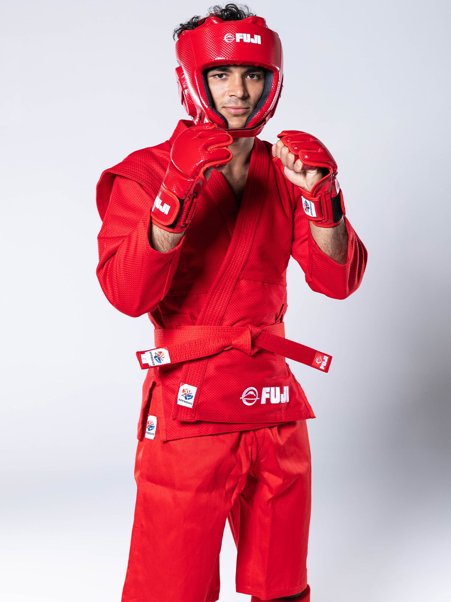 A person wearing bright red FIAS Sambo Combat Gloves, a matching red martial arts uniform, and a red helmet stands in a fighting stance against a plain background.