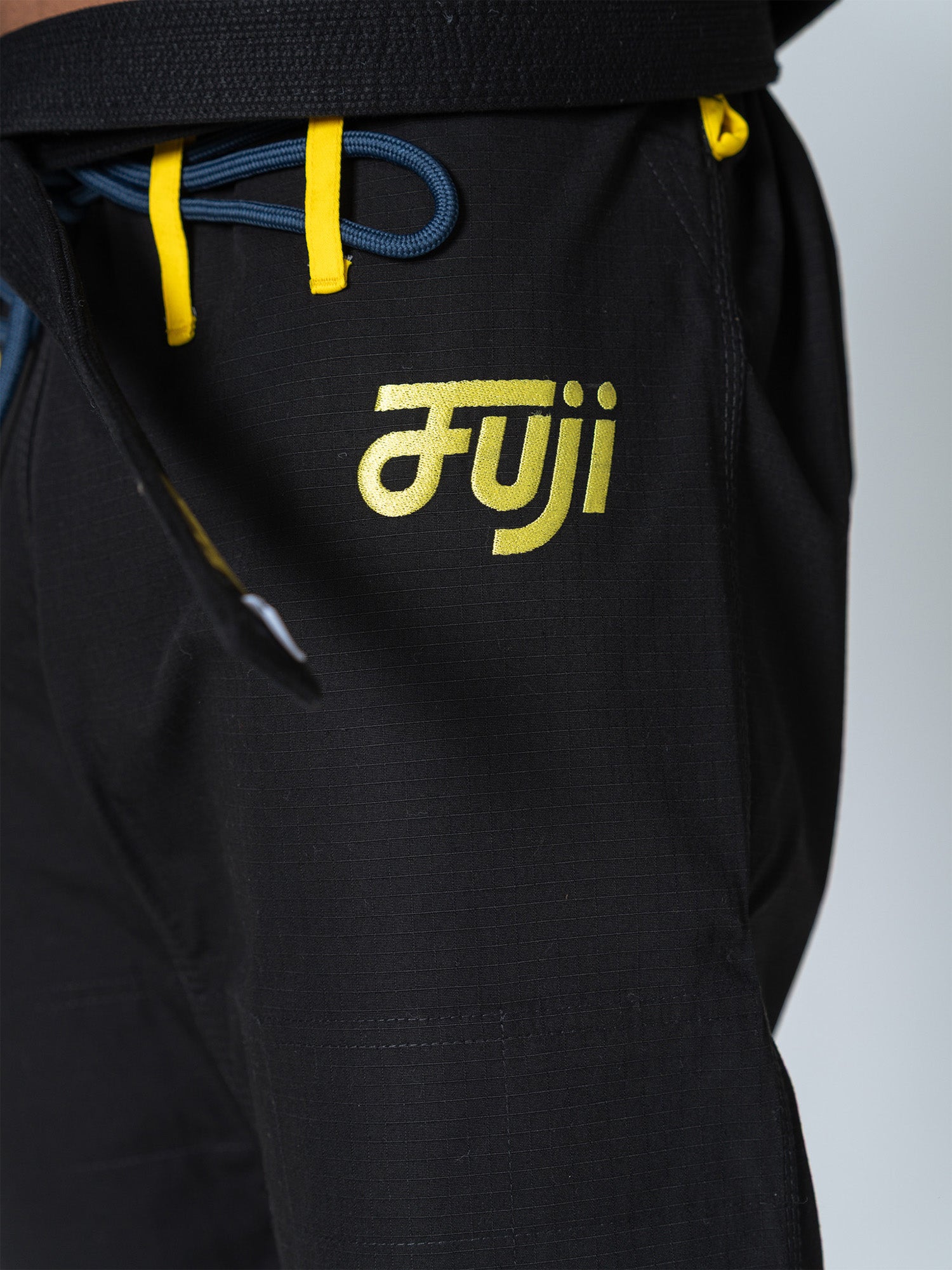 Close-up of the Special Edition Holiday Flow Tech Jiu Jitsu Gi in black, featuring yellow and blue drawstrings. "Fuji" is printed in yellow on the left thigh. The textured fabric looks durable.
