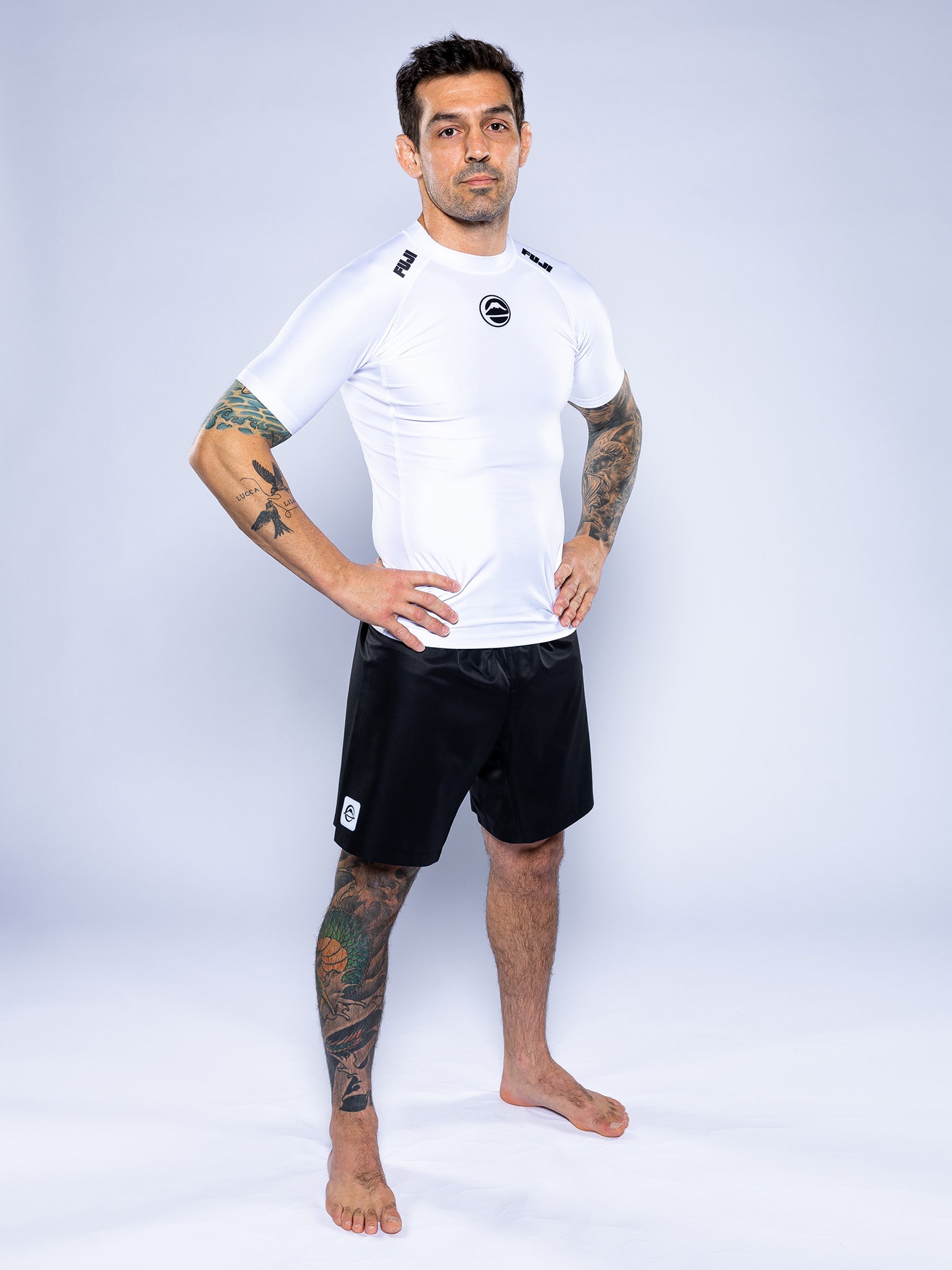 A man with tattoos stands barefoot on a white background, wearing the Baseline 2.0 Short Sleeve Rashguard in white and black shorts, with his hands on his hips.