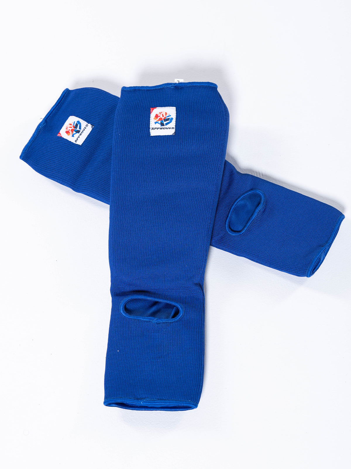 Two FIAS Sambo Combat Shin Guards in blue, featuring heel openings and small rectangular patches with a red, blue, and white logo near the top edge, are crossed on a white background.