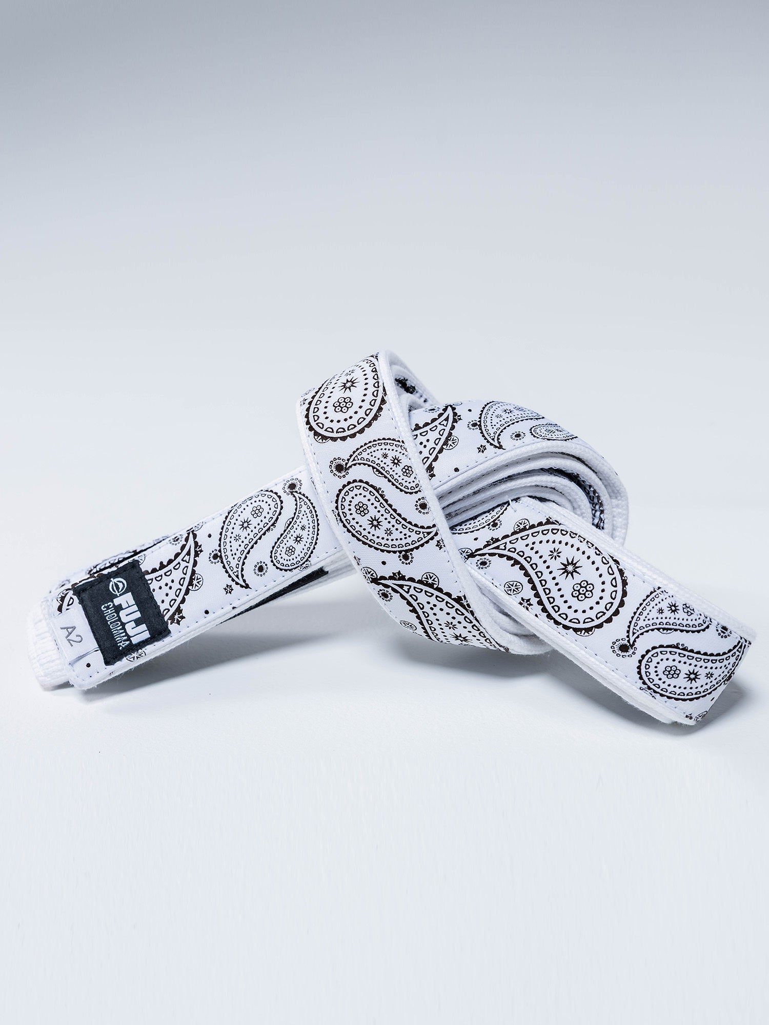 The CholoMMA Paisley Pearl Weave BJJ Belt White, featuring a black paisley pattern and a black label with white text, is coiled on a light surface.