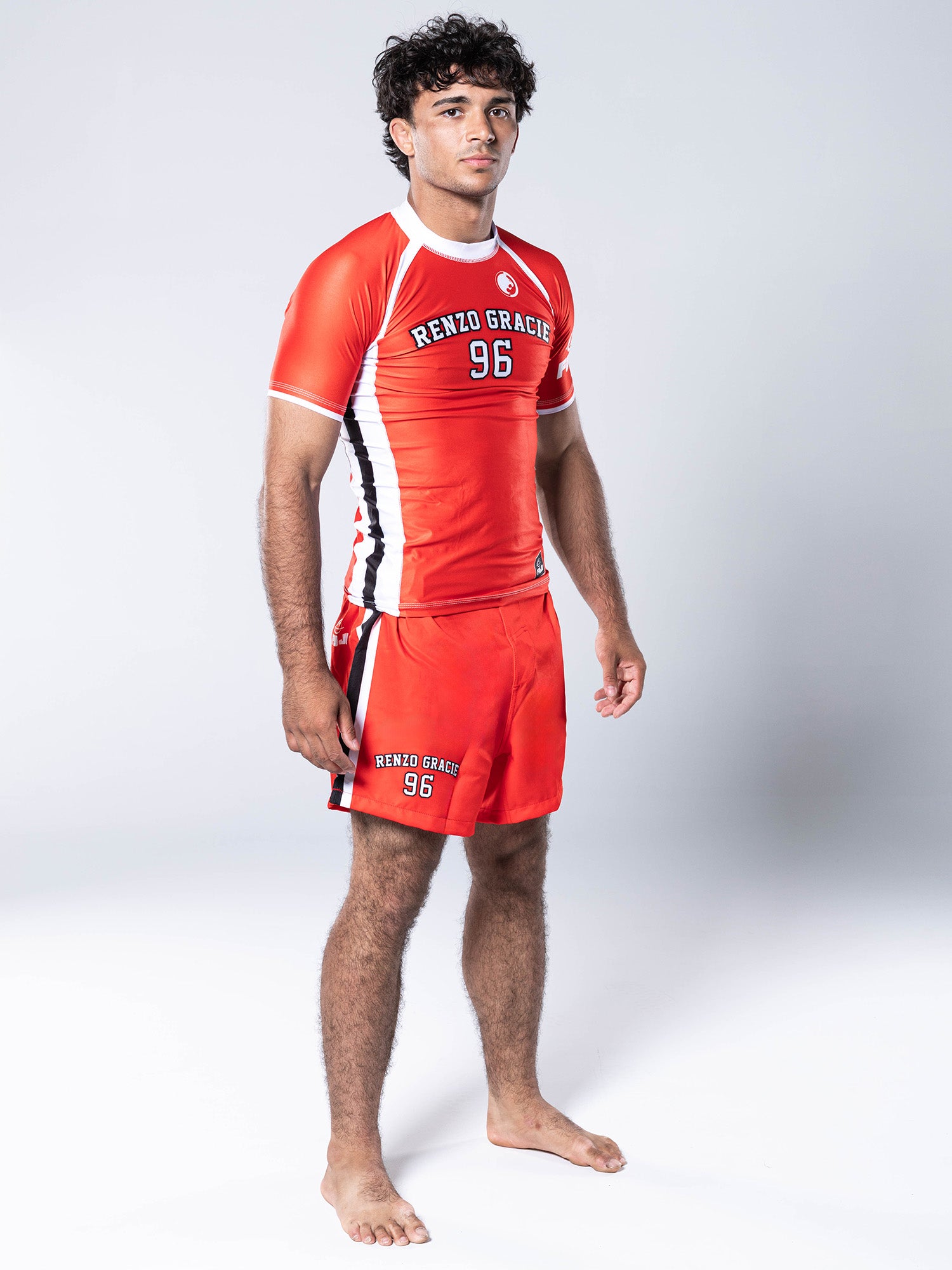 A young man stands barefoot on a white background, wearing the Renzo Gracie '96 Jersey Short Sleeve Rashguard in red along with matching shorts, looking forward with a neutral expression.