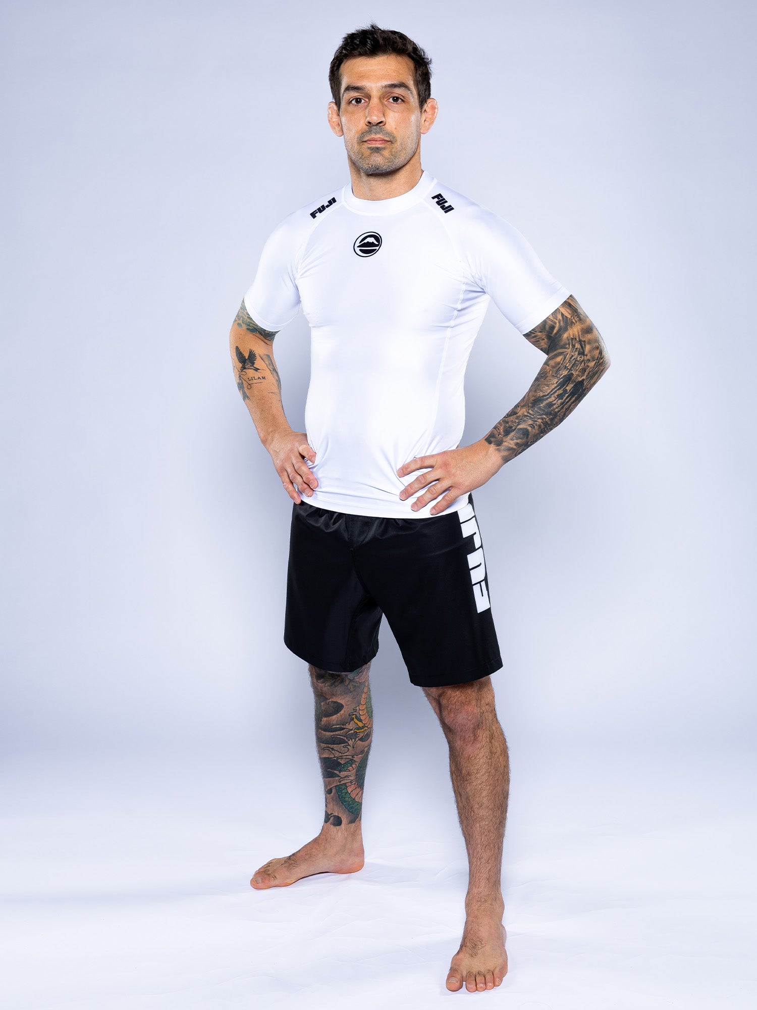 A tattooed man stands barefoot on a white background, confidently posing with hands on hips while wearing the Baseline 2.0 Short Sleeve Rashguard White and black grappling shorts with white text, looking at the camera.