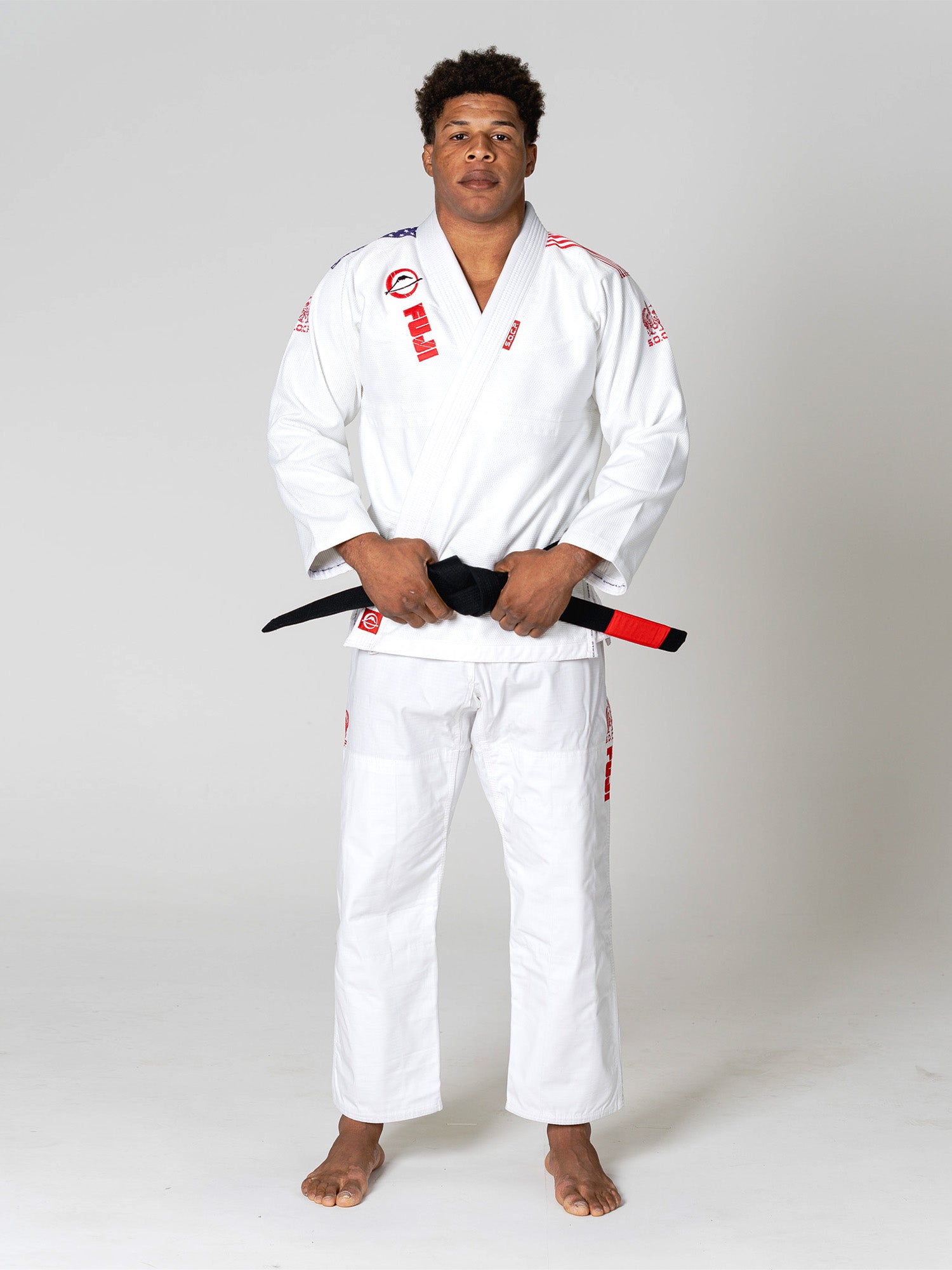 A man wearing the S.O.C.P. Sekai BJJ Gi 2.0 White with red patches stands barefoot on a white background, holding a black belt with a red bar in front of him.