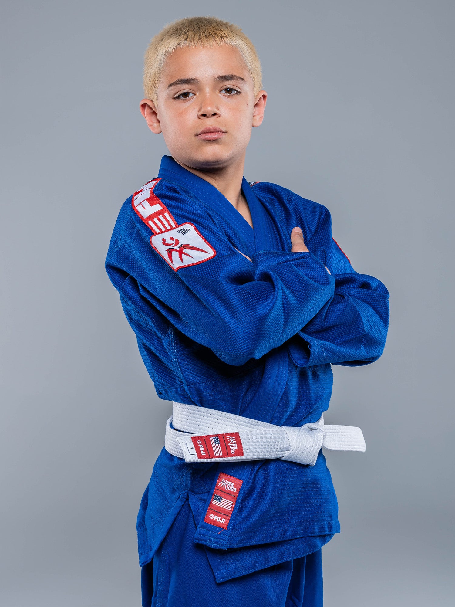 A young boy with short blond hair stands confidently with arms crossed, wearing a USA Judo Single Weave Gi 2.0 Blue and a white belt against a plain gray background.