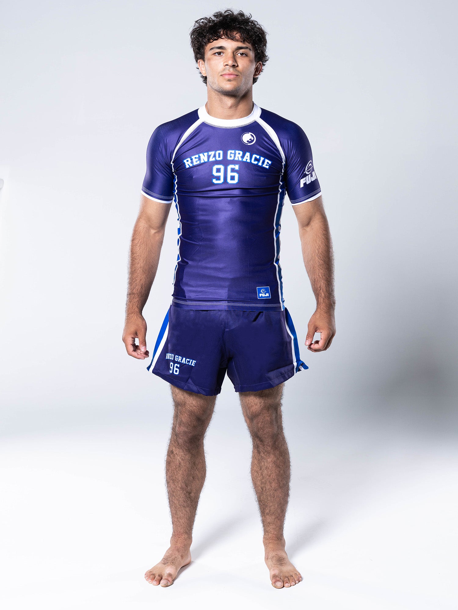 A man stands barefoot on a white background, facing forward with his arms relaxed at his sides, wearing the Renzo Gracie '96 Jersey Short Sleeve Rashguard in blue and matching shorts.