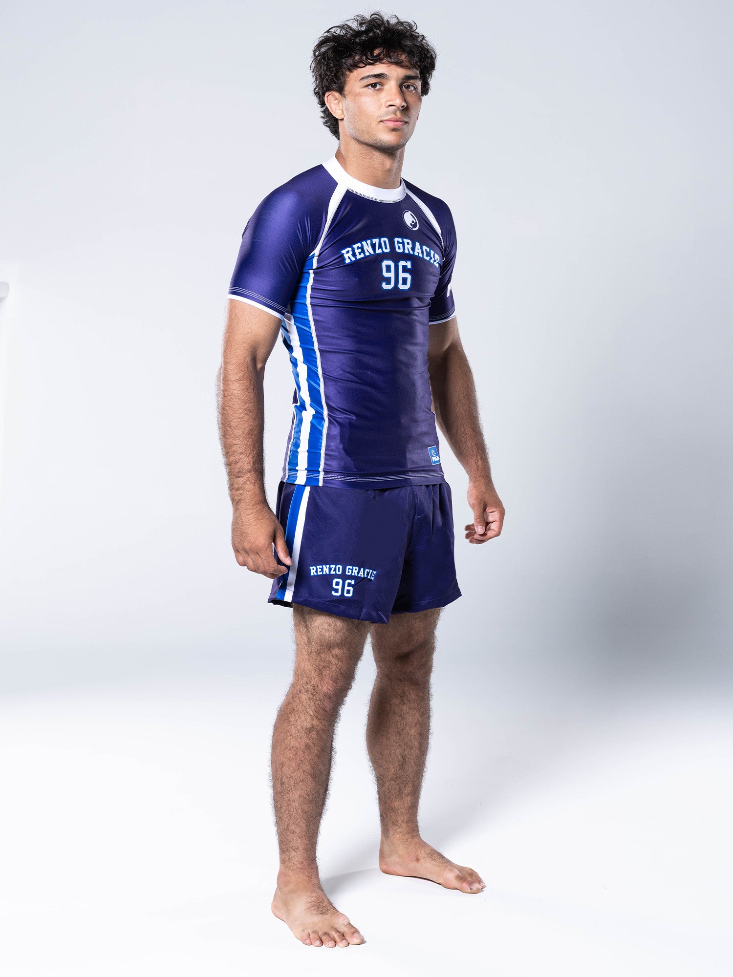 A man in a blue Renzo Gracie '96 Jersey Short Sleeve Rashguard and matching shorts stands barefoot on a white background, facing forward with a neutral expression.