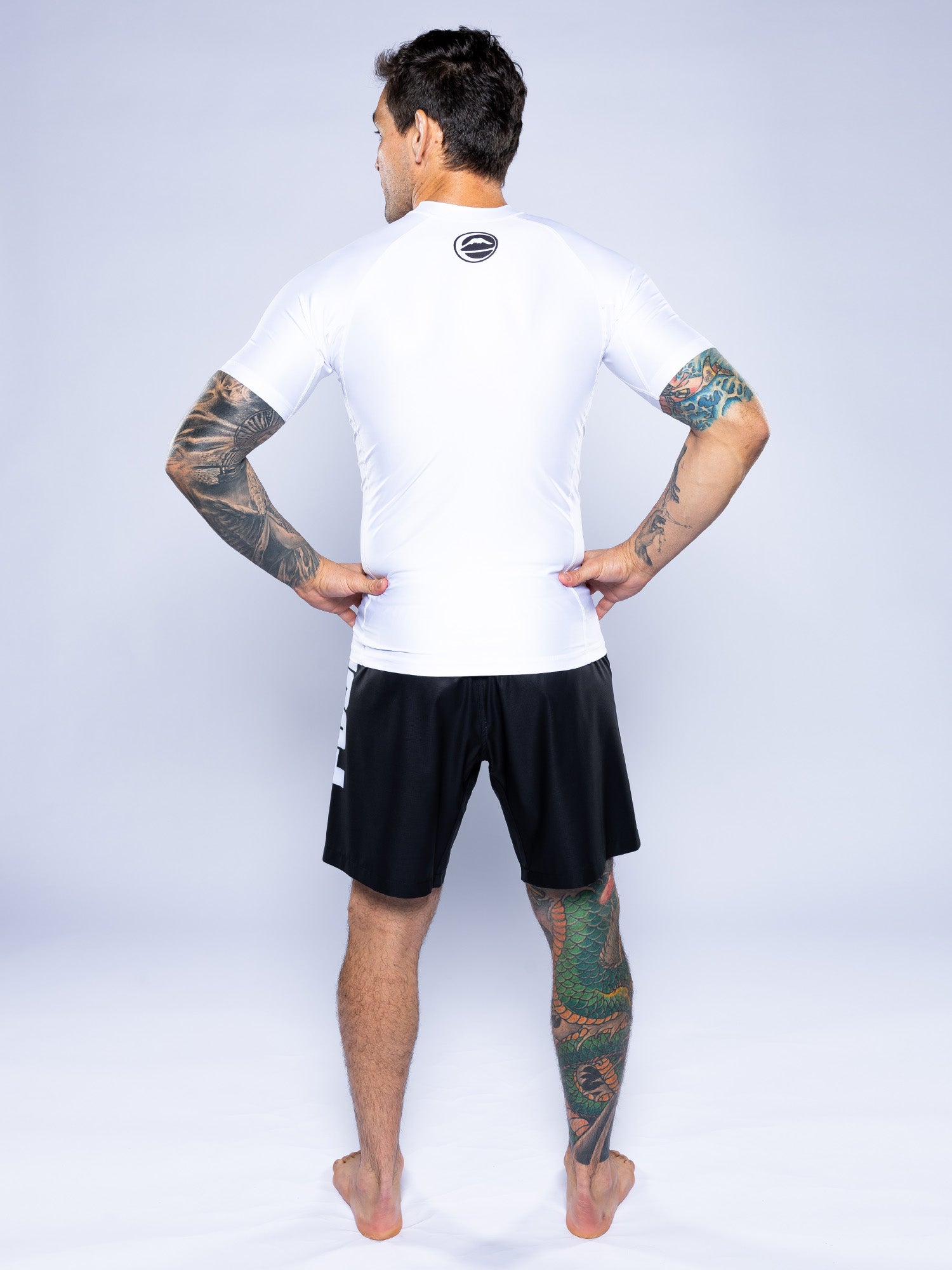 A barefoot man stands facing away, hands on hips, wearing the Baseline 2.0 Short Sleeve Rashguard White displaying a back logo and black shorts. Colorful tattoos cover his arms and one leg. The background is plain and light-colored.