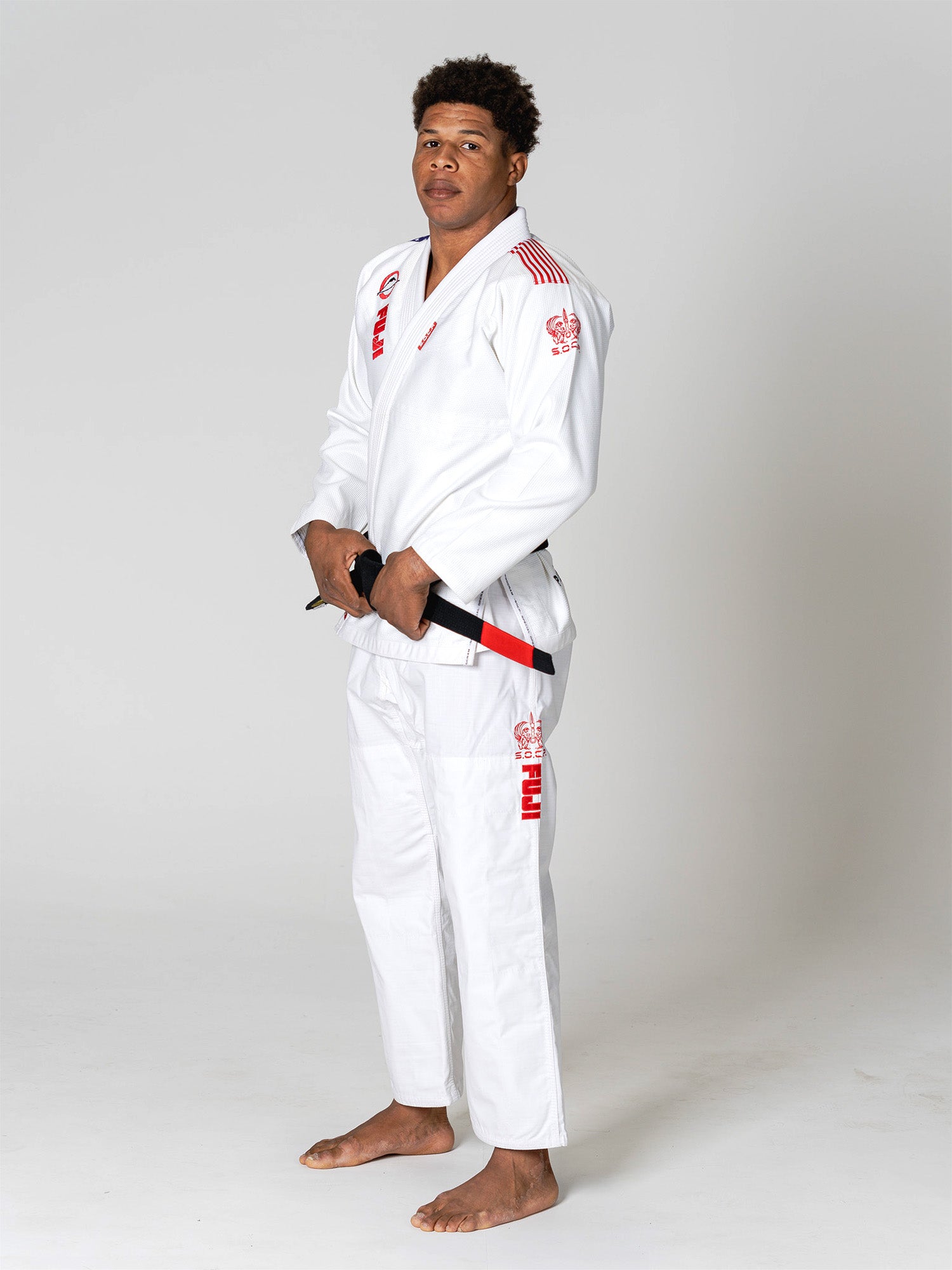 A man stands barefoot on a white background, wearing the S.O.C.P. Sekai BJJ Gi 2.0 White with red details. He confidently holds a black belt with both hands and looks toward the camera.