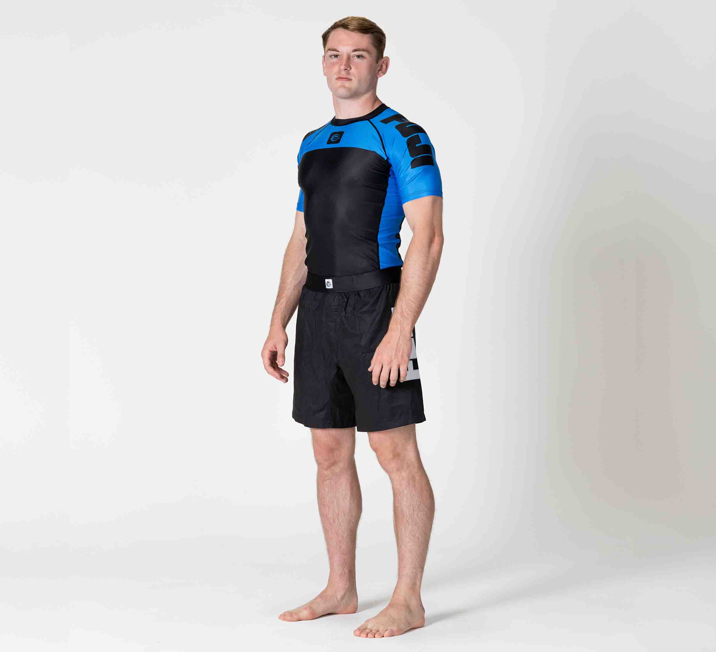 A man stands barefoot on a white background, wearing the Competition Ranked Rashguard Blue and black shorts, with a neutral expression and his arms by his sides.