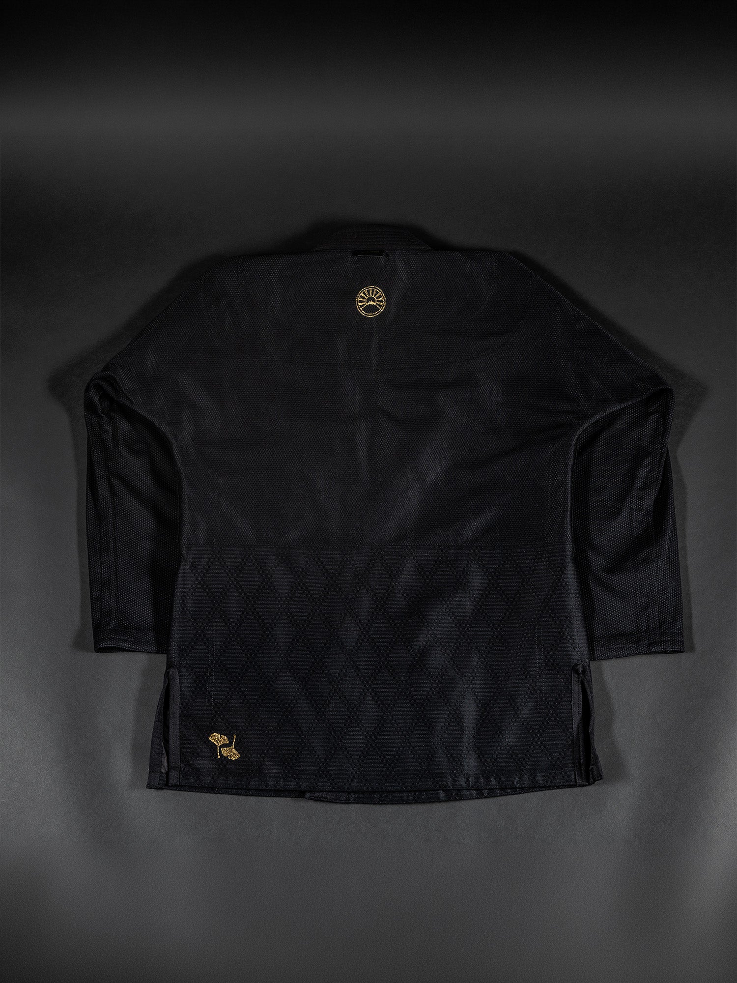 A Heritage Keisho Lightweight BJJ Gi in black is laid flat, showing the back view with subtle diamond patterns across the lower half and small gold embroidered logos near the collar and bottom corner.