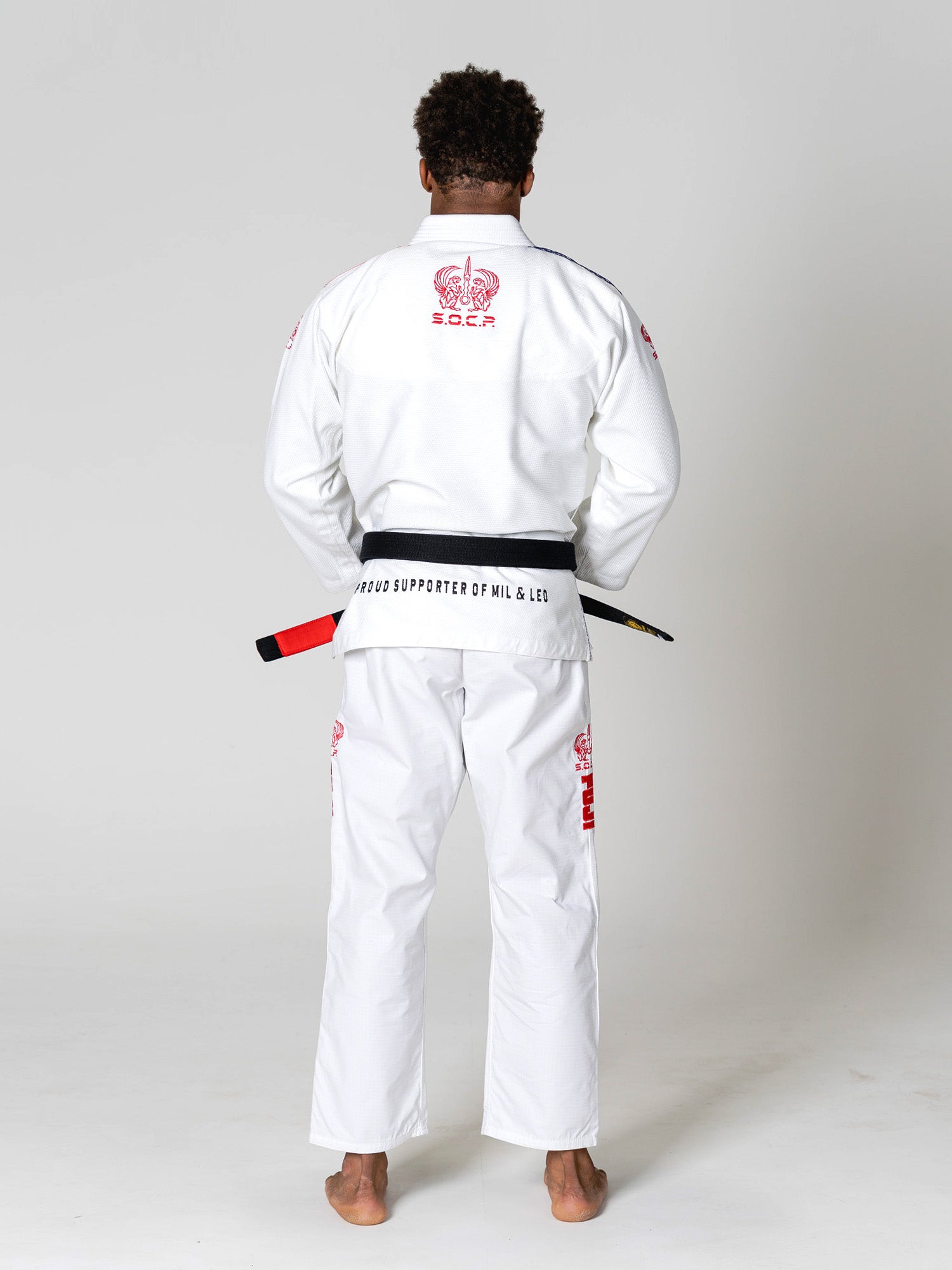 A barefoot person in a white S.O.C.P. Sekai BJJ Gi 2.0 with red embroidery stands facing away, holding a black and red belt behind their back against a plain light background.