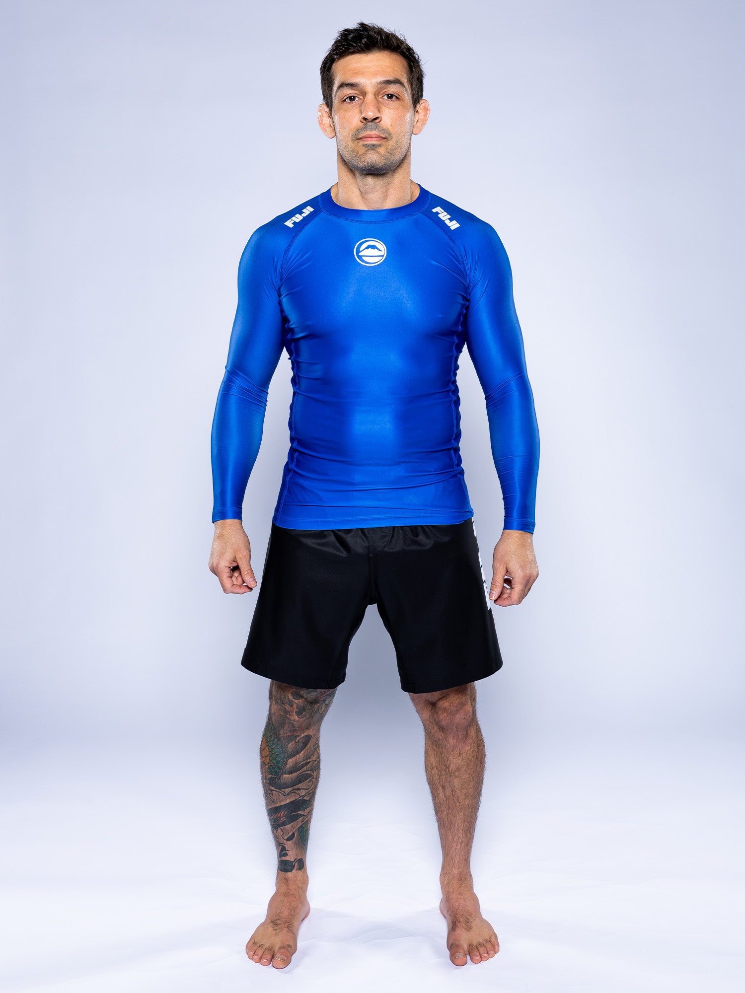 A man with short dark hair and tattoos on his left leg stands barefoot against a white background, wearing the Baseline 2.0 Long Sleeve Rashguard Blue and black shorts, with a neutral expression.