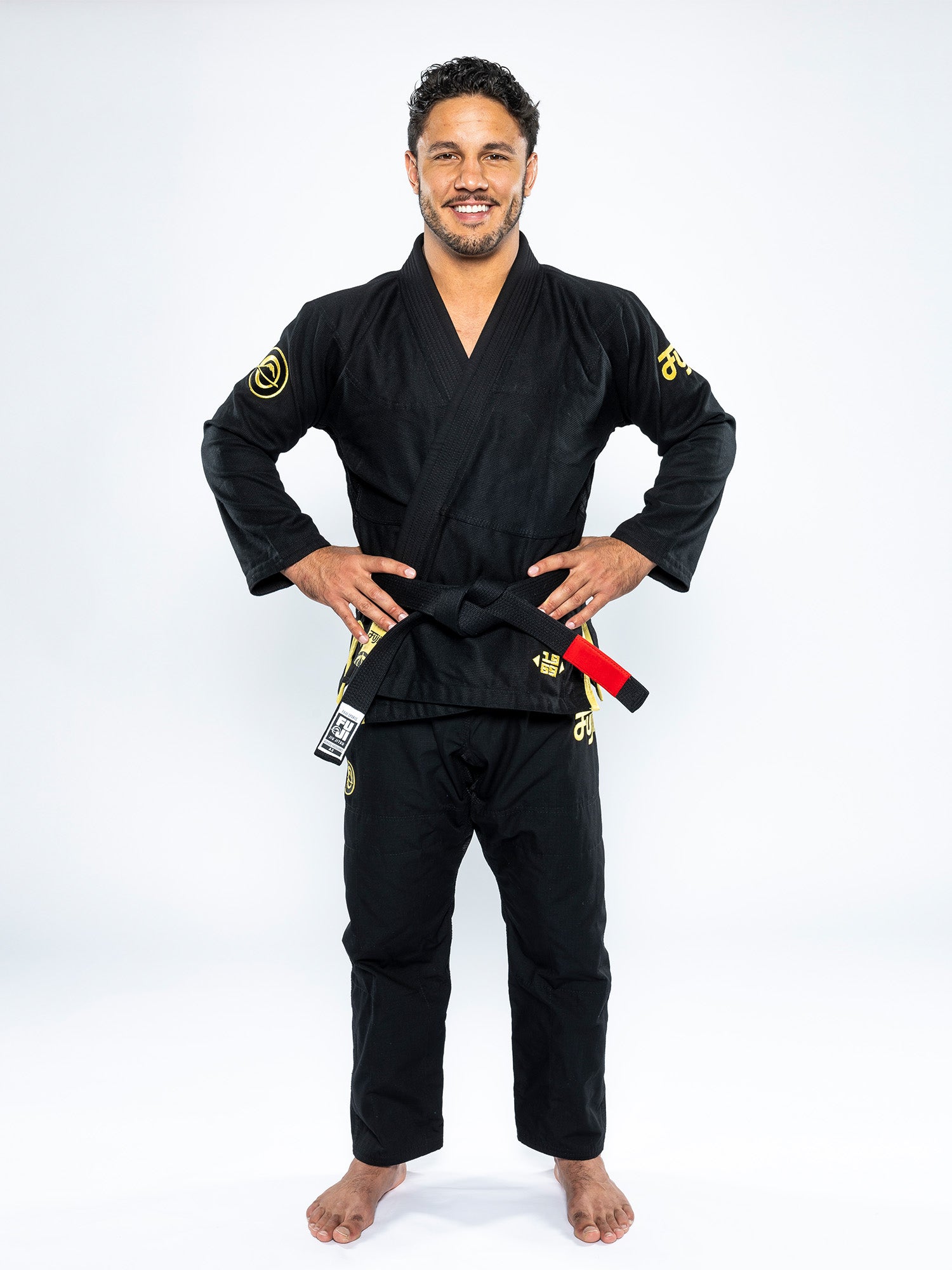 A man stands barefoot on a white background, smiling with his hands on his hips. He is wearing the Flow-Tech BJJ Gi Black/Gold with yellow accents and a black belt featuring a red stripe.