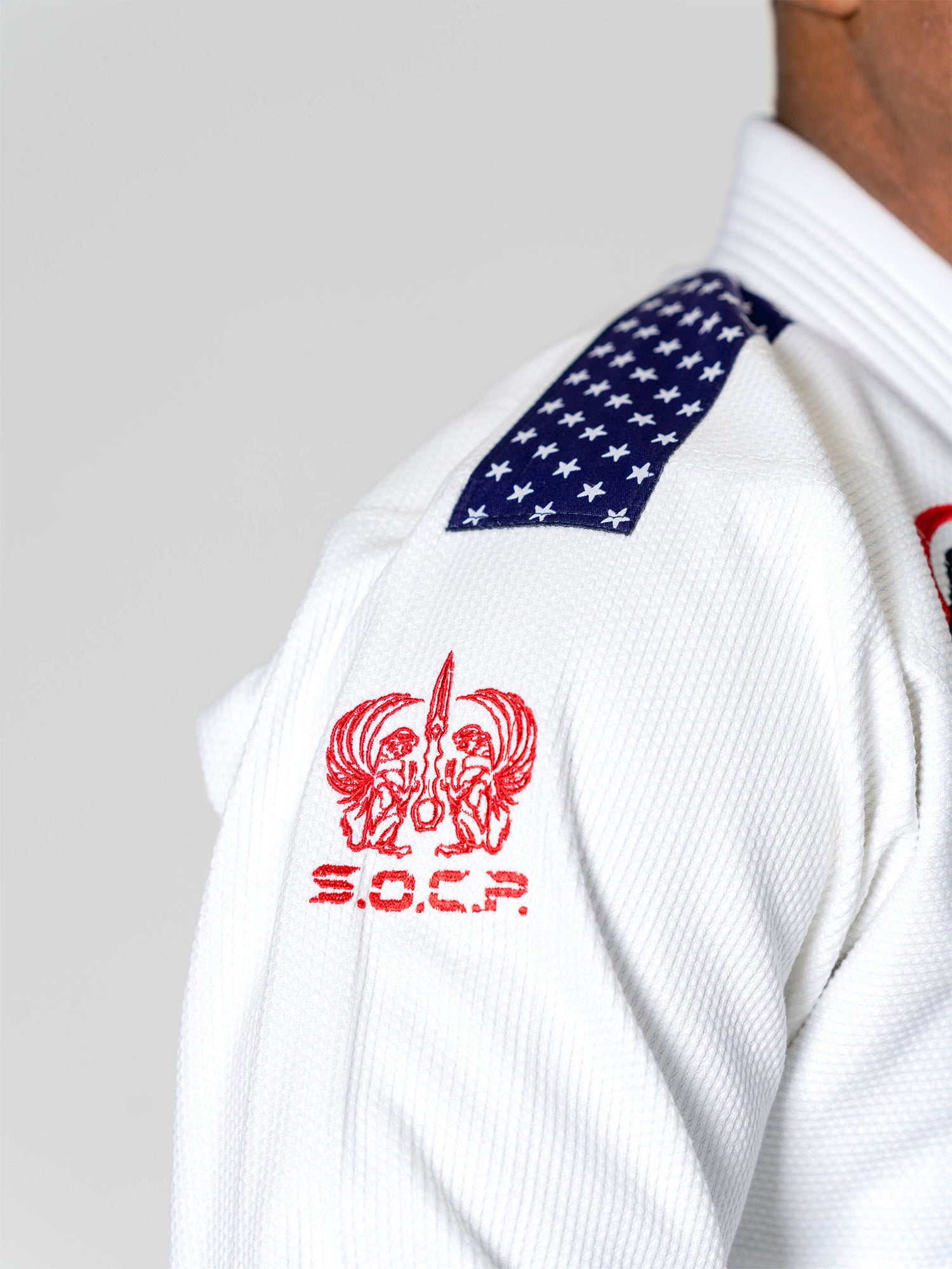 Close-up of a person wearing the S.O.C.P. Sekai BJJ Gi 2.0 White, featuring a red embroidered logo with two lion heads and S.O.C.P. lettering on the sleeve, and a shoulder patch with white stars on a blue background.