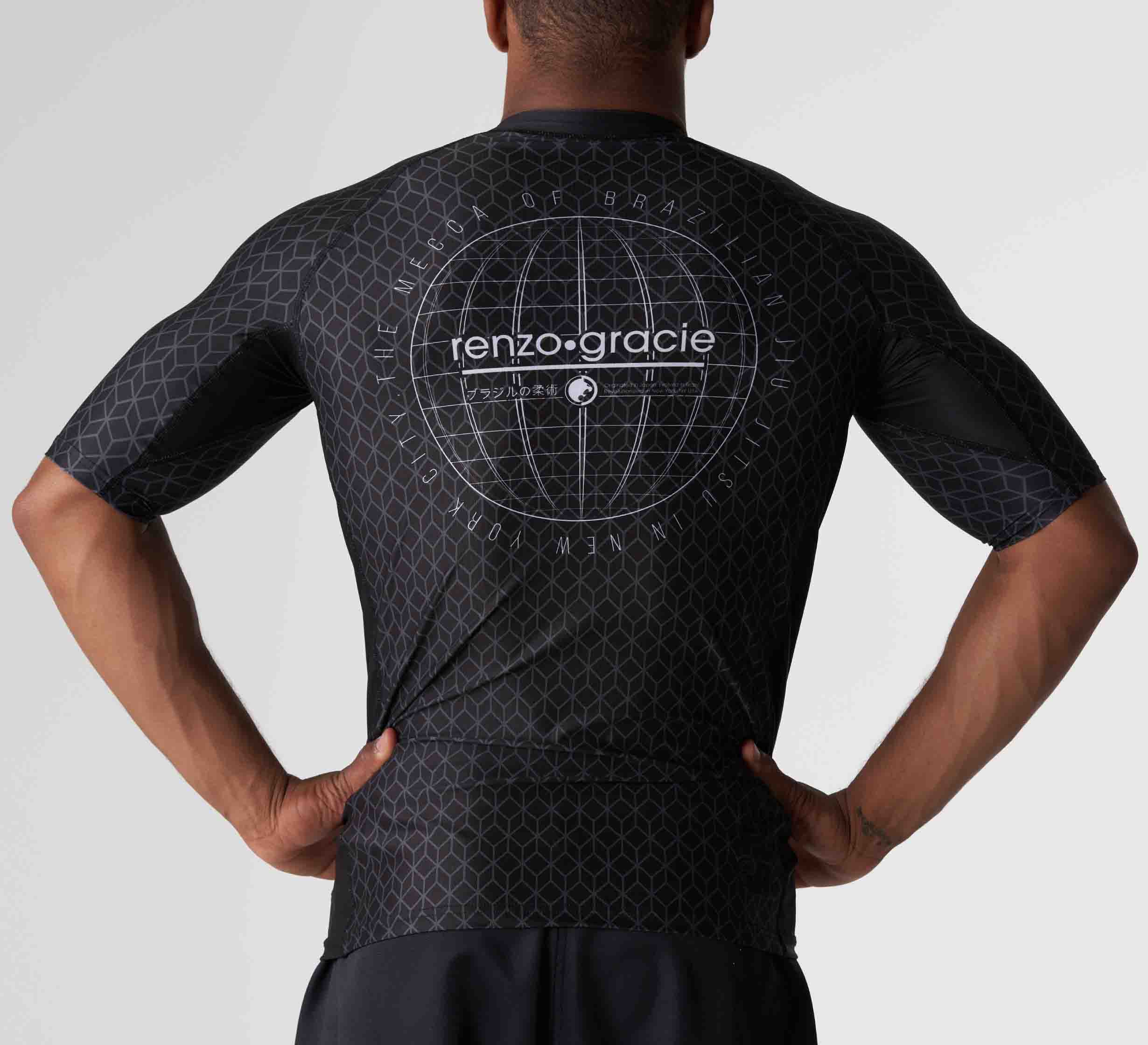 A person stands with hands on hips, wearing the Renzo Gracie Mecca Ranked Short Sleeve Rashguard featuring a black geometric pattern and renzo.gracie with a globe graphic printed on the back.