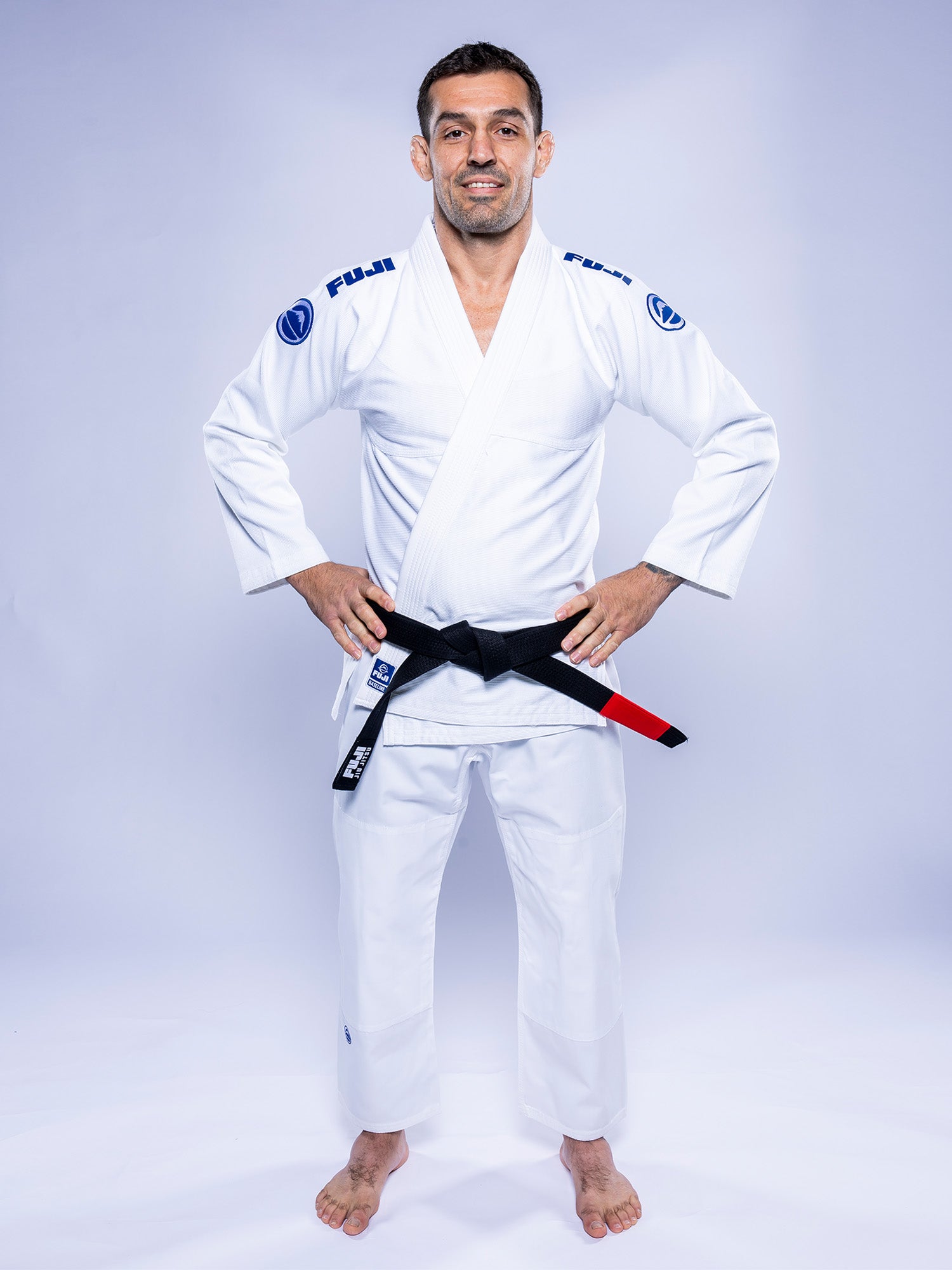 A man smiles with hands on hips, barefoot on a white floor, wearing the Baseline 2.0 BJJ Gi White Navy and a black belt against a plain light background.