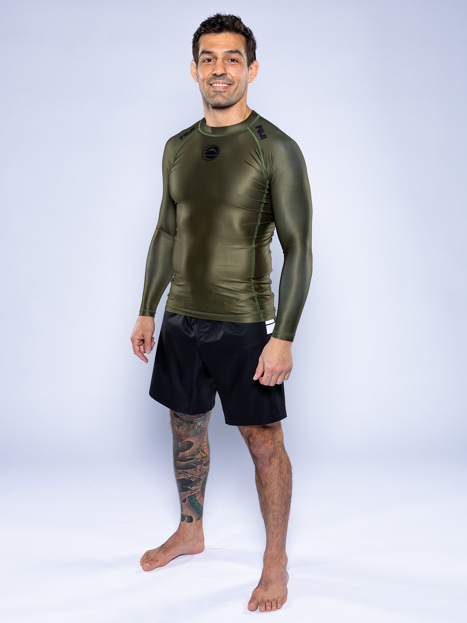 A man with short dark hair stands barefoot on a light floor, smiling at the camera. He wears the Baseline 2.0 Long Sleeve Rashguard in Military Green and black shorts, showing a large tattoo on his lower right leg.