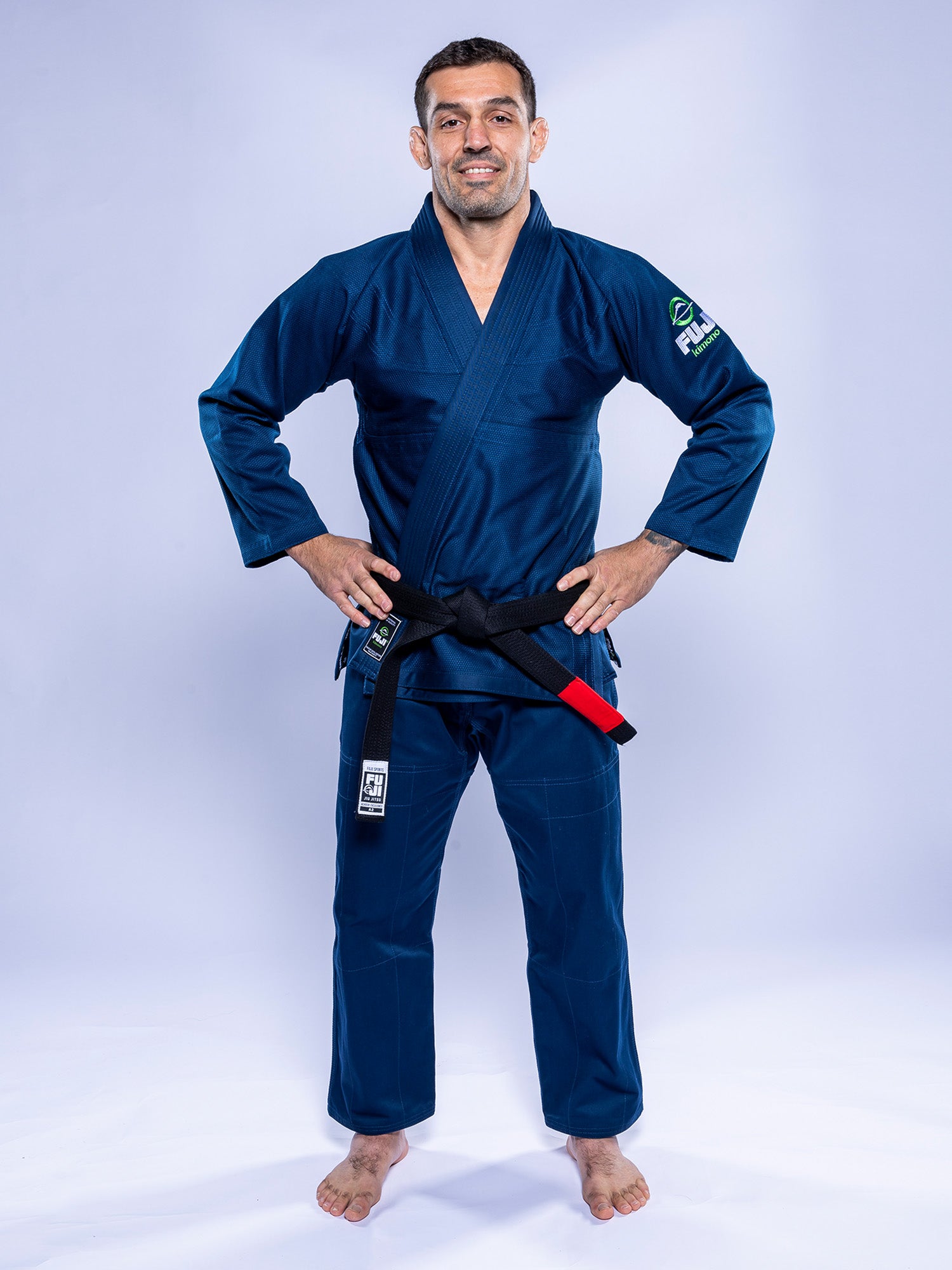 A man wearing the Lightweight BJJ Gi Navy and a black belt stands barefoot against a light background, smiling with his hands on his hips.