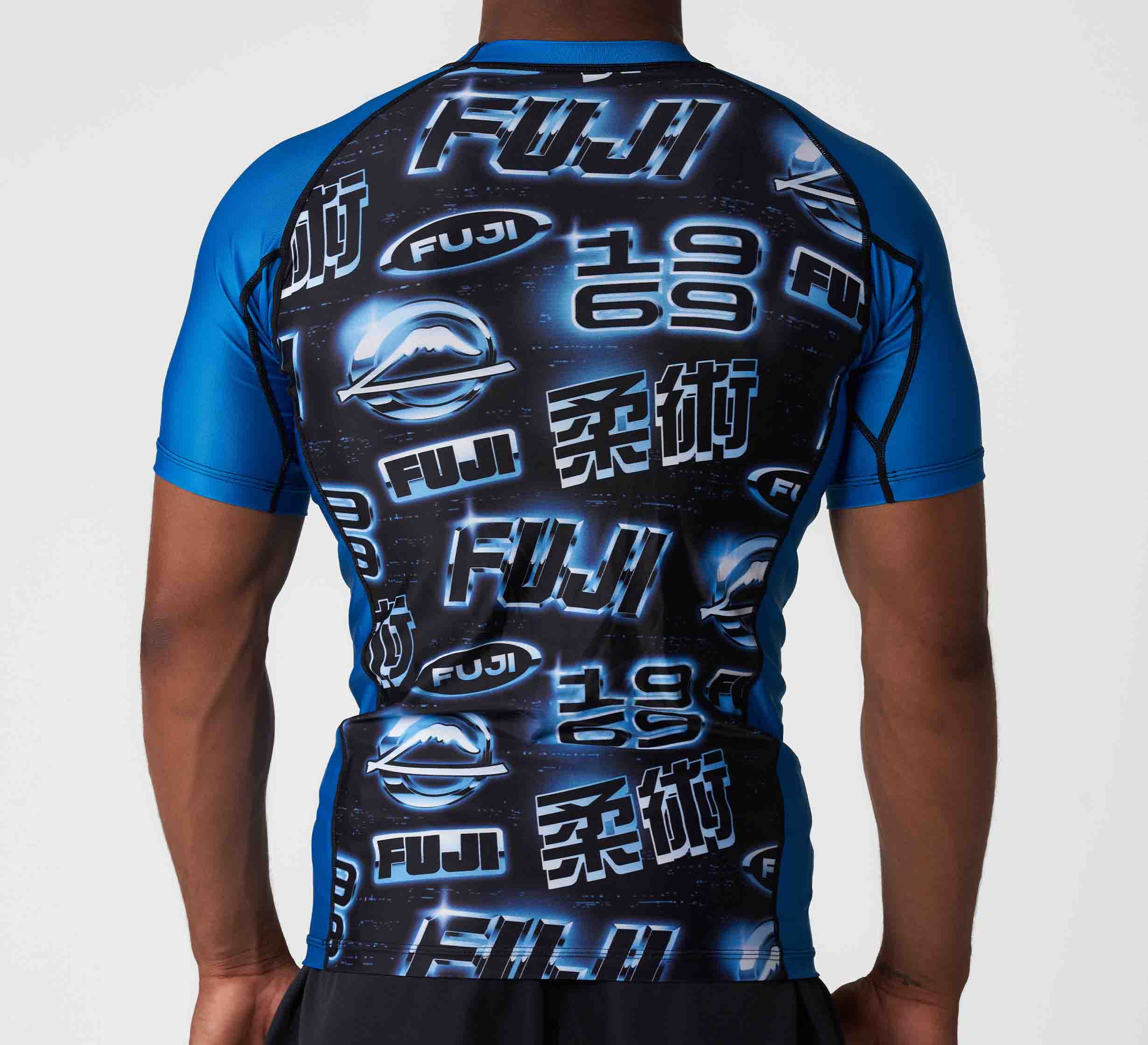 A person seen from the back wearing the Synth Flex Lite Rashguard Blue, a blue and black athletic shirt with bold FUJI graphics, "1969," and various white and blue logos on a dark, space-themed background.